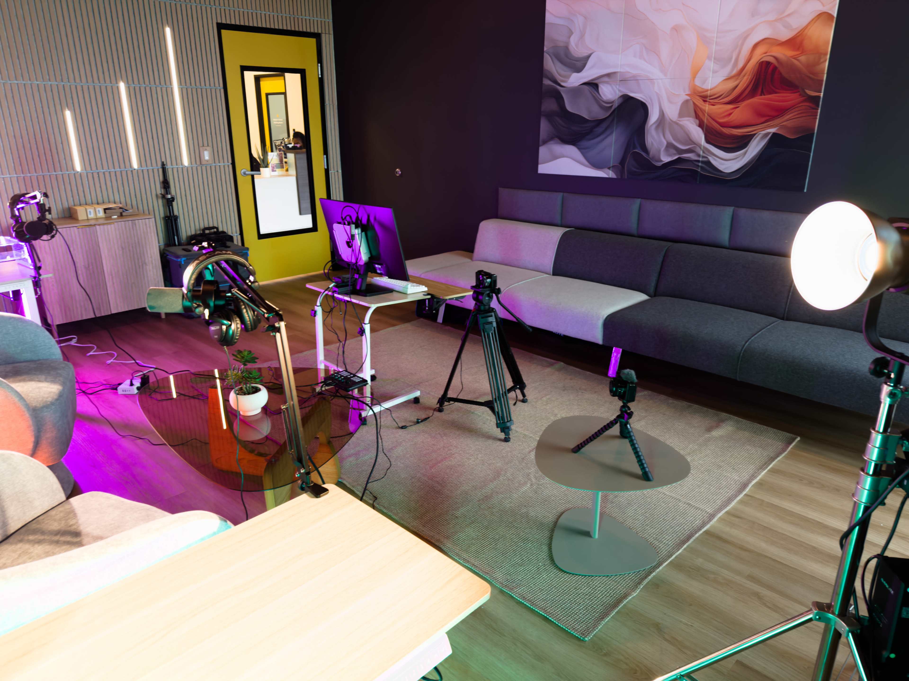 The image shows a modern living room setup with a gray sofa, a computer workstation with cameras and lighting equipment, and colorful decorative elements.