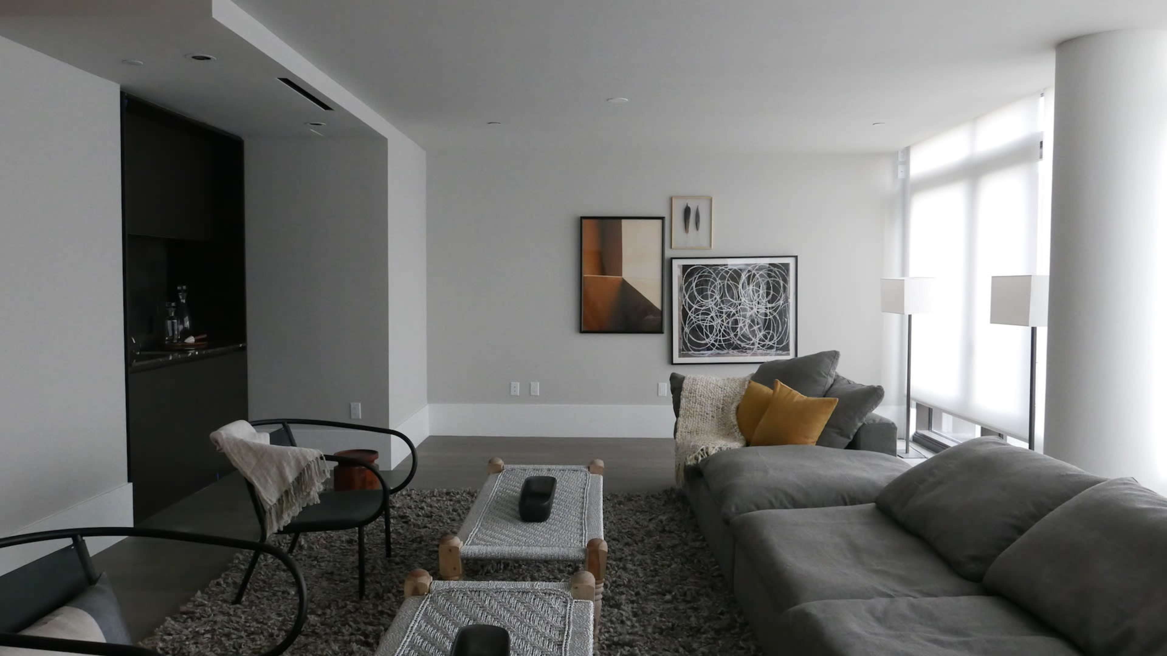A modern living room features a gray sectional sofa, a textured rug, and a coffee table, with artwork hanging on the wall and natural light streaming through large windows.