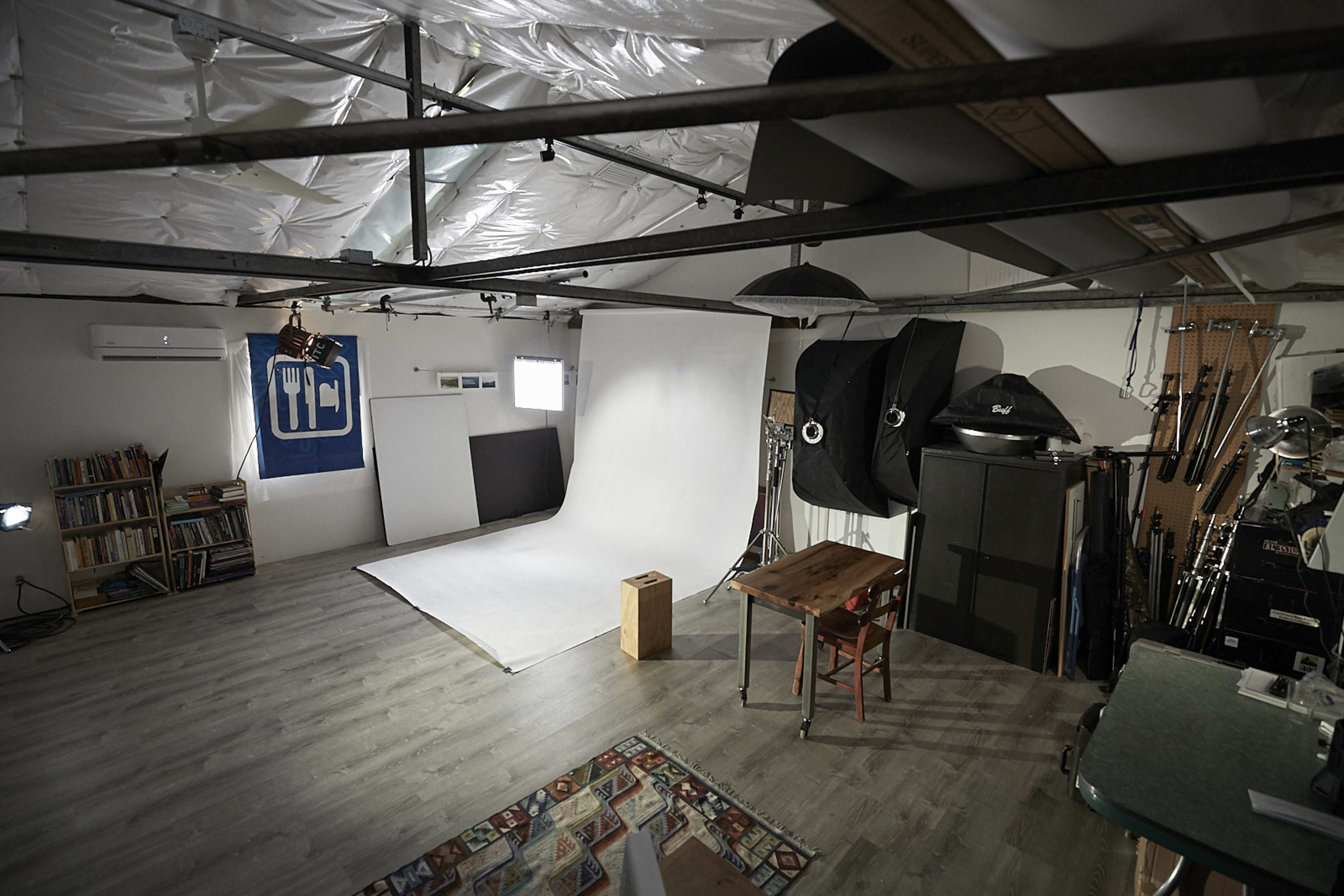 The image shows a photography studio with a white backdrop, a wooden table and chair, and various props and equipment on the walls.