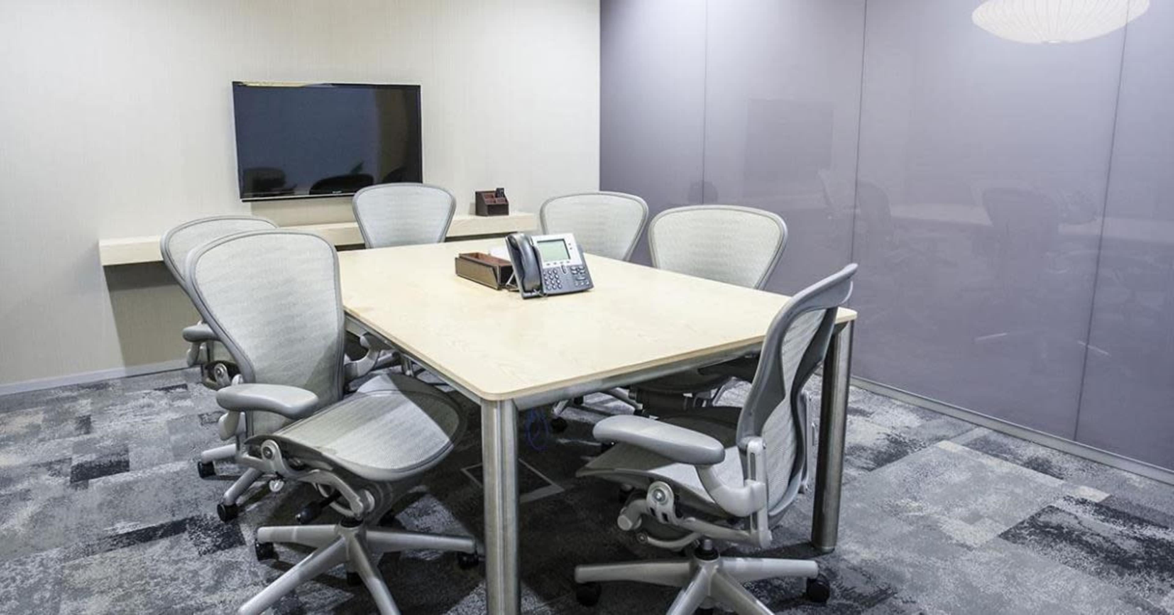 A conference room features a rectangular table surrounded by six ergonomic chairs, with a television on the wall and a phone at the center of the table.