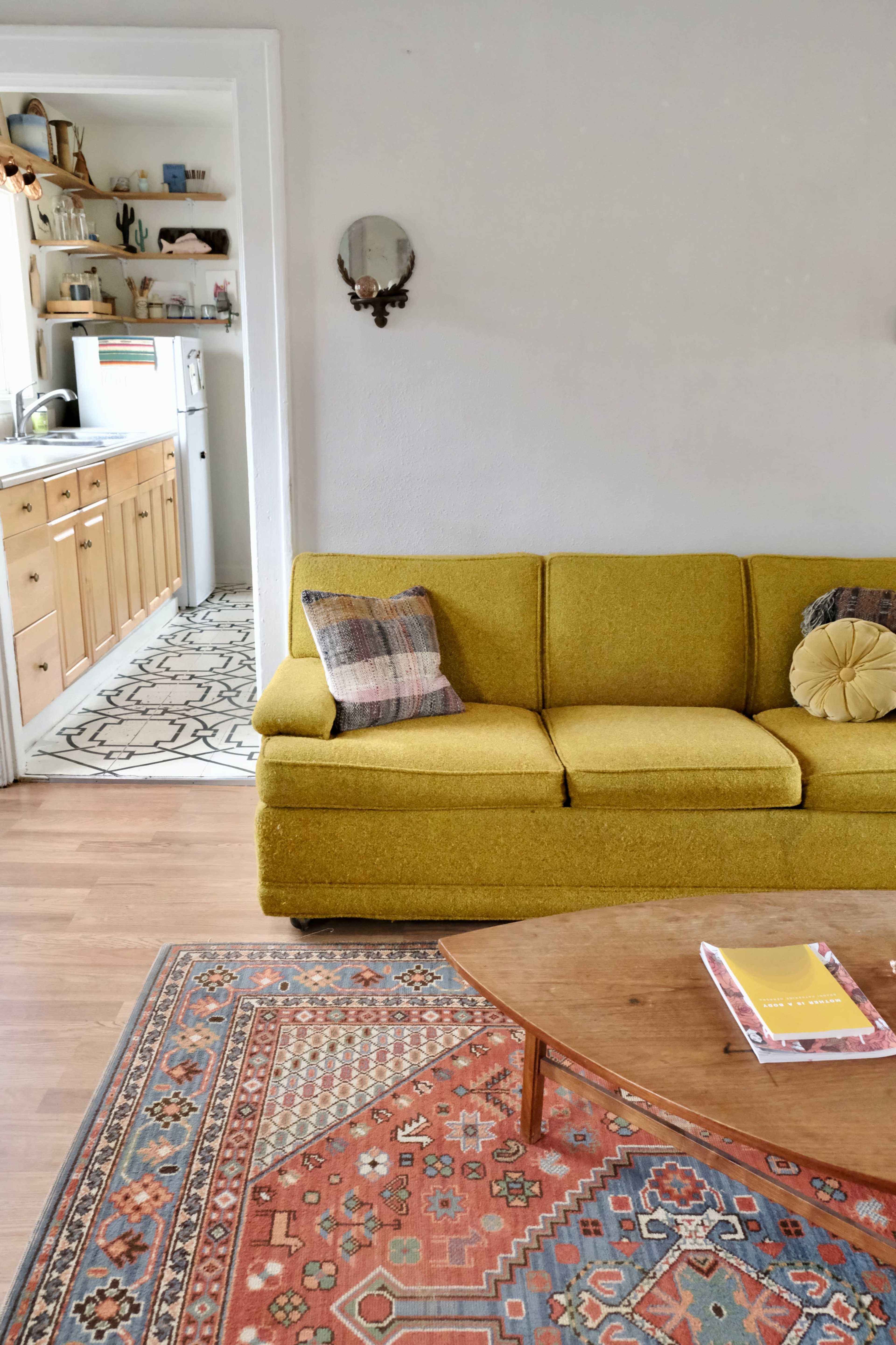 A cozy living room with a yellow sofa, a patterned rug, and a view into a kitchen area in the background.