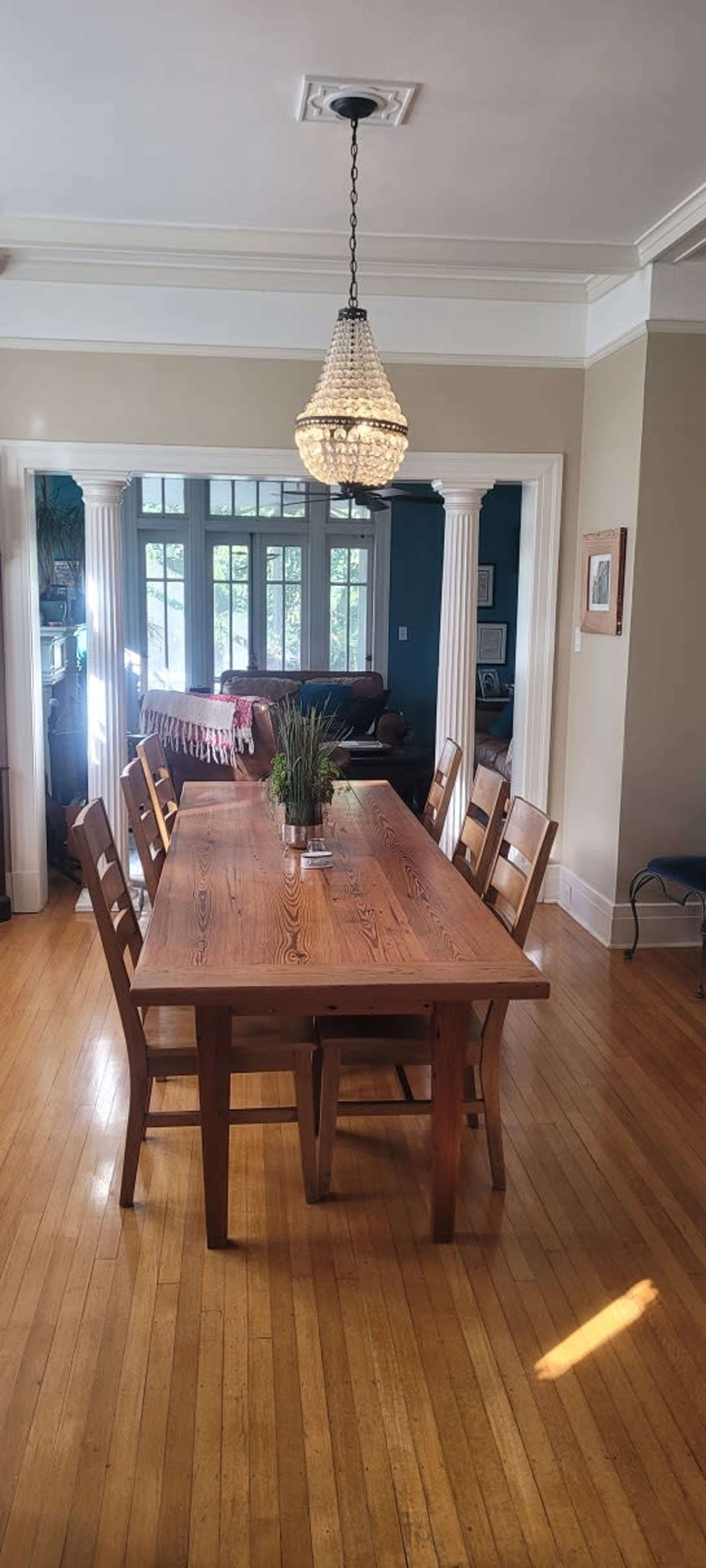 A long wooden dining table with eight chairs is illuminated by a chandelier, set in a brightly lit room featuring large windows and wooden flooring.