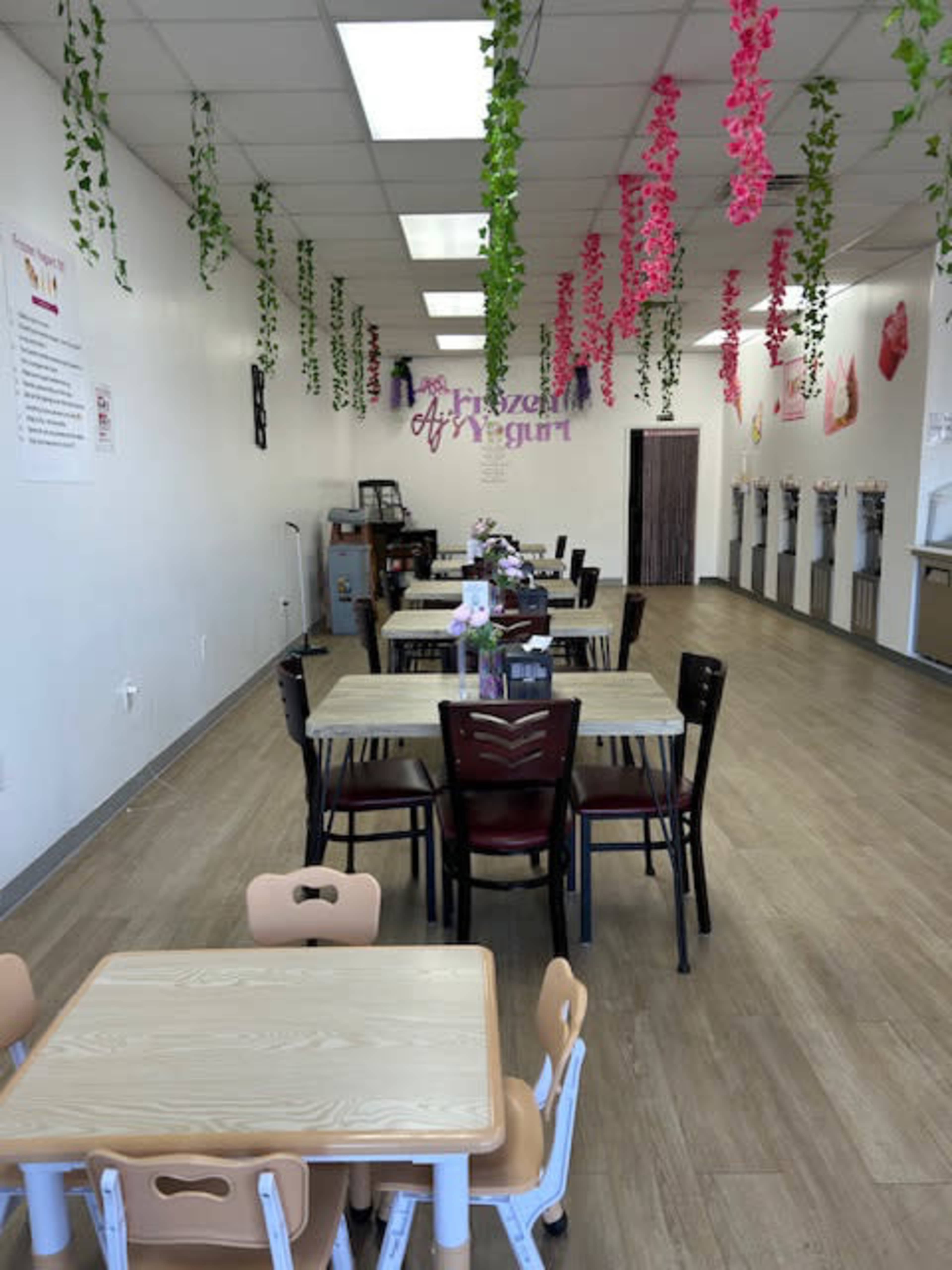 The image shows a brightly lit yogurt shop featuring several tables and chairs, with decorative hanging plants and an area for self-serve yogurt dispensers.