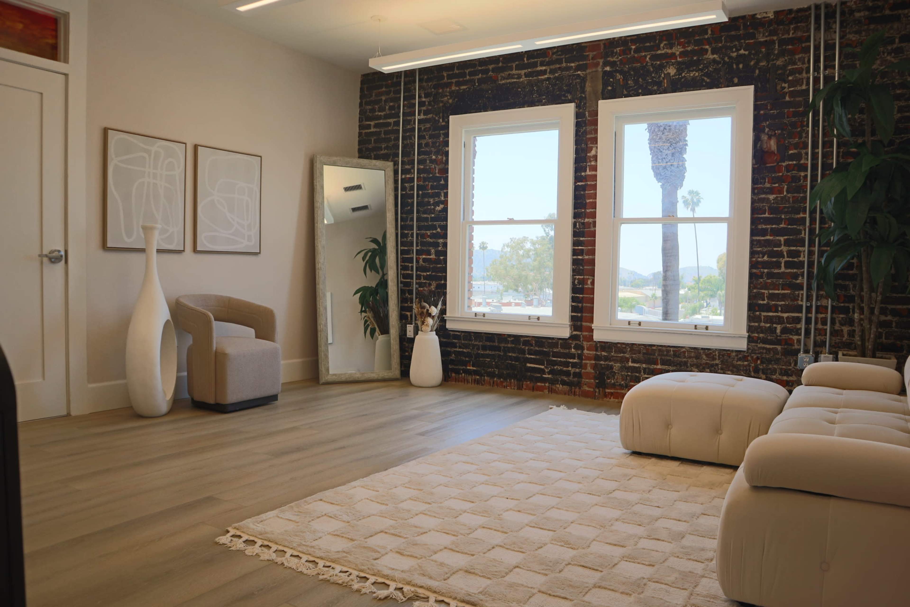 The room features exposed brick walls, large windows, and minimalist furniture, including a chair and two poufs on a textured rug.