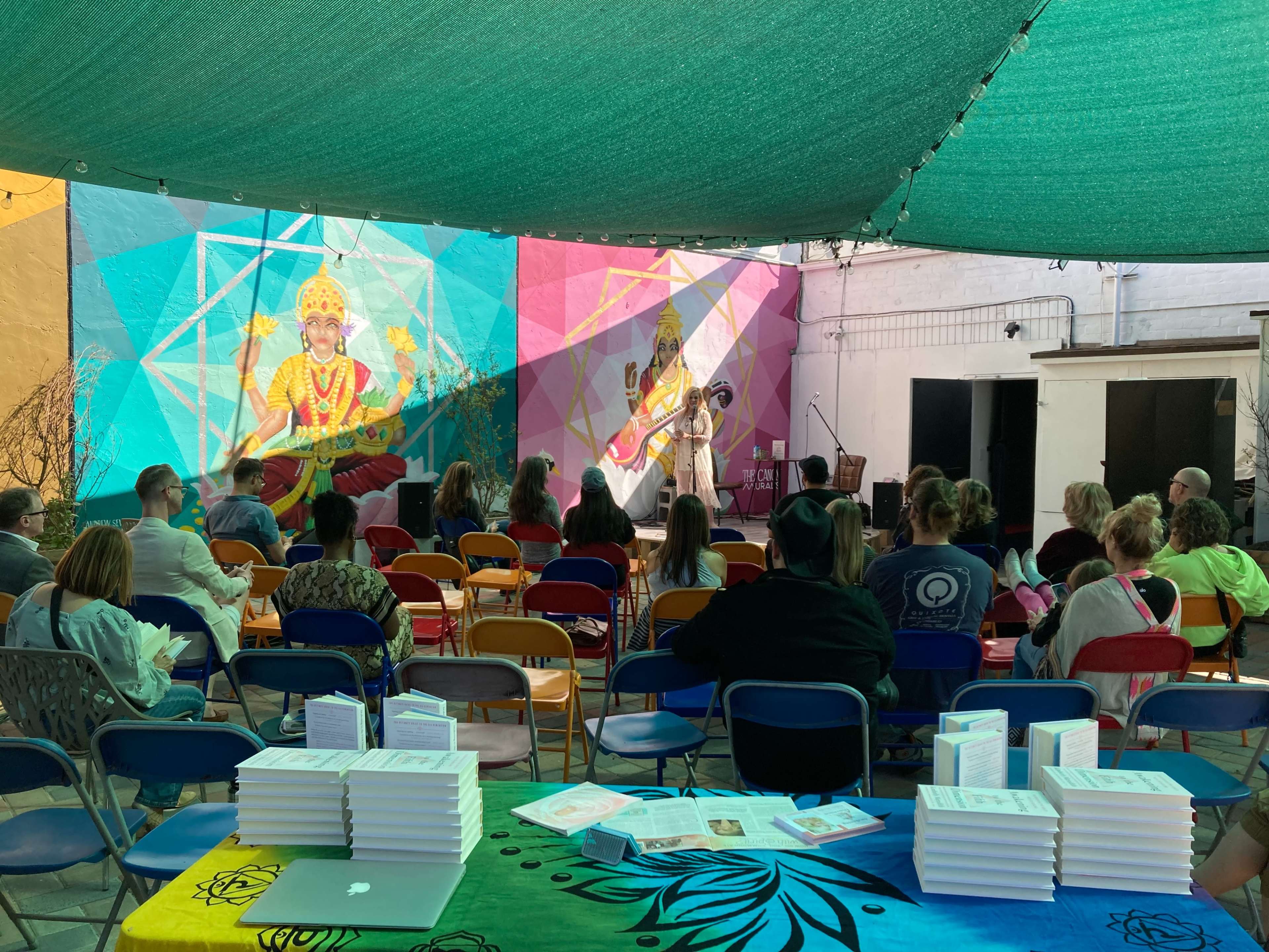 A crowd gathers under a colorful canopy in an outdoor space, facing a large mural depicting a musician.