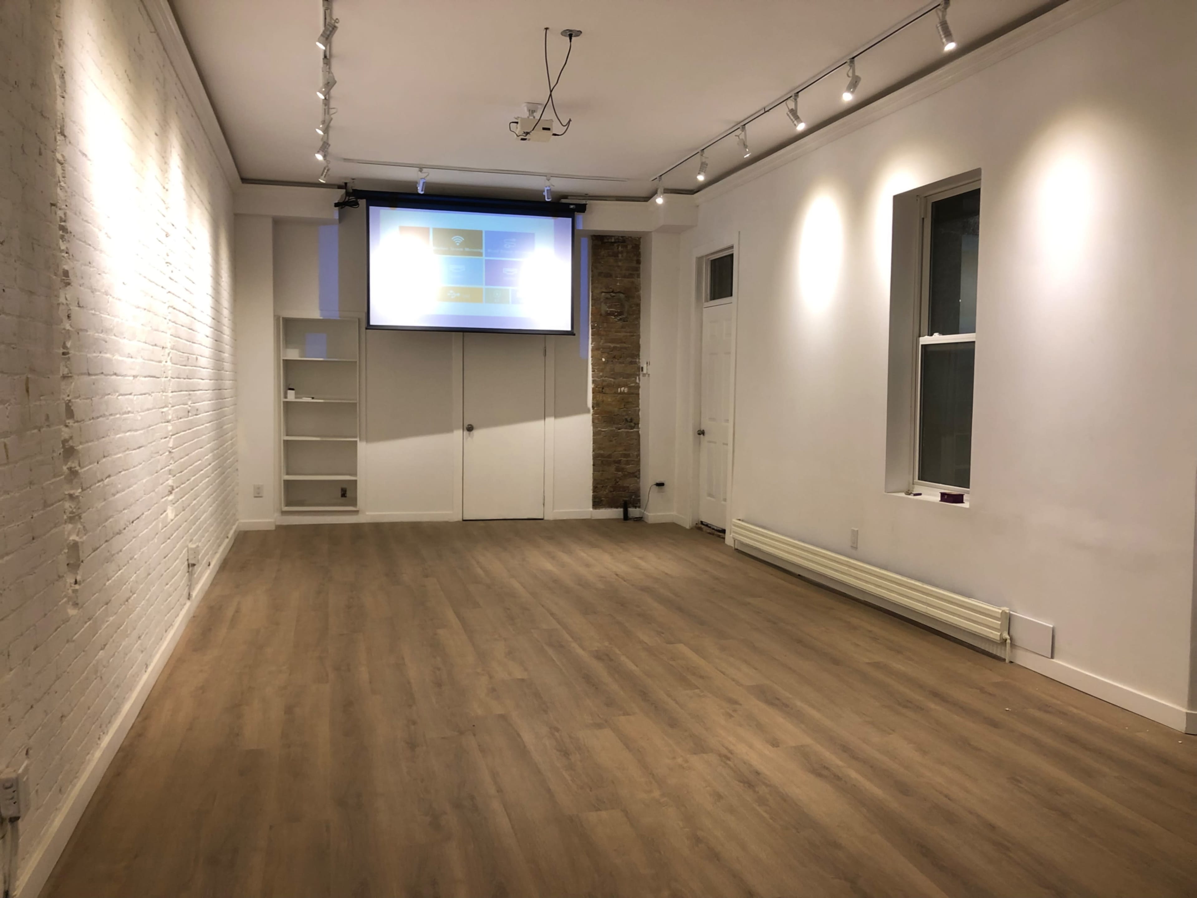 A spacious, empty room with light-colored walls, a wooden floor, and a projector screen on the front wall.