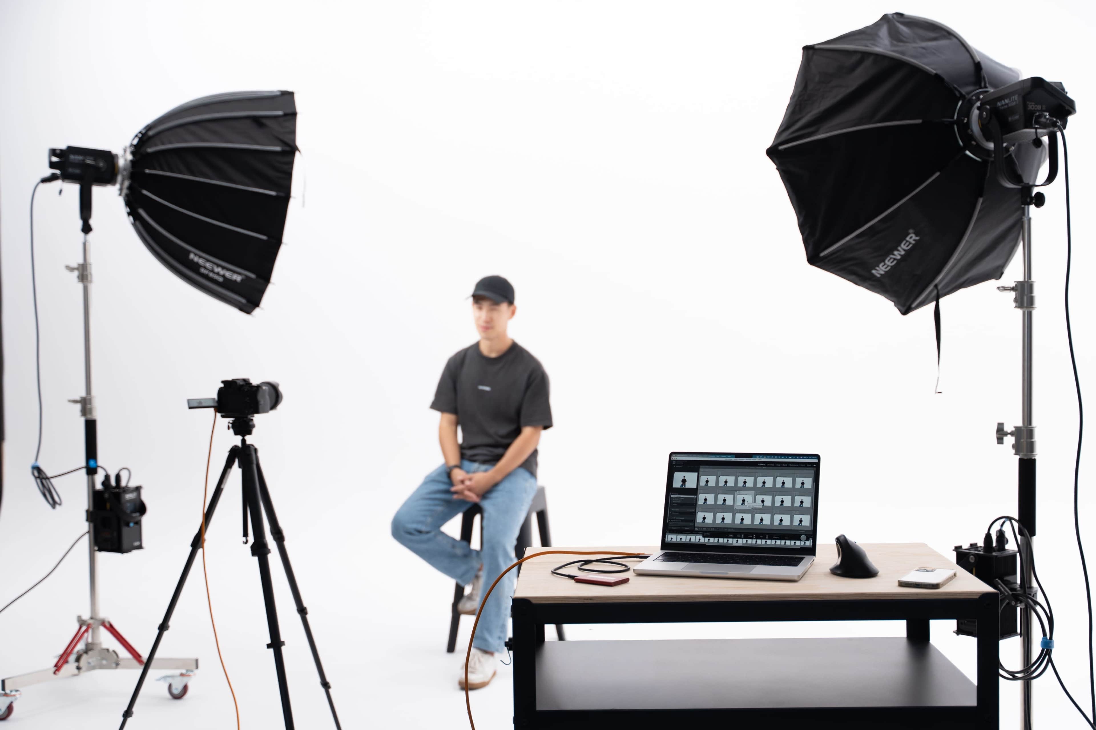 A man sits on a stool in a studio setup with two softbox lights, a camera on a tripod, and a laptop displaying images on a table.
