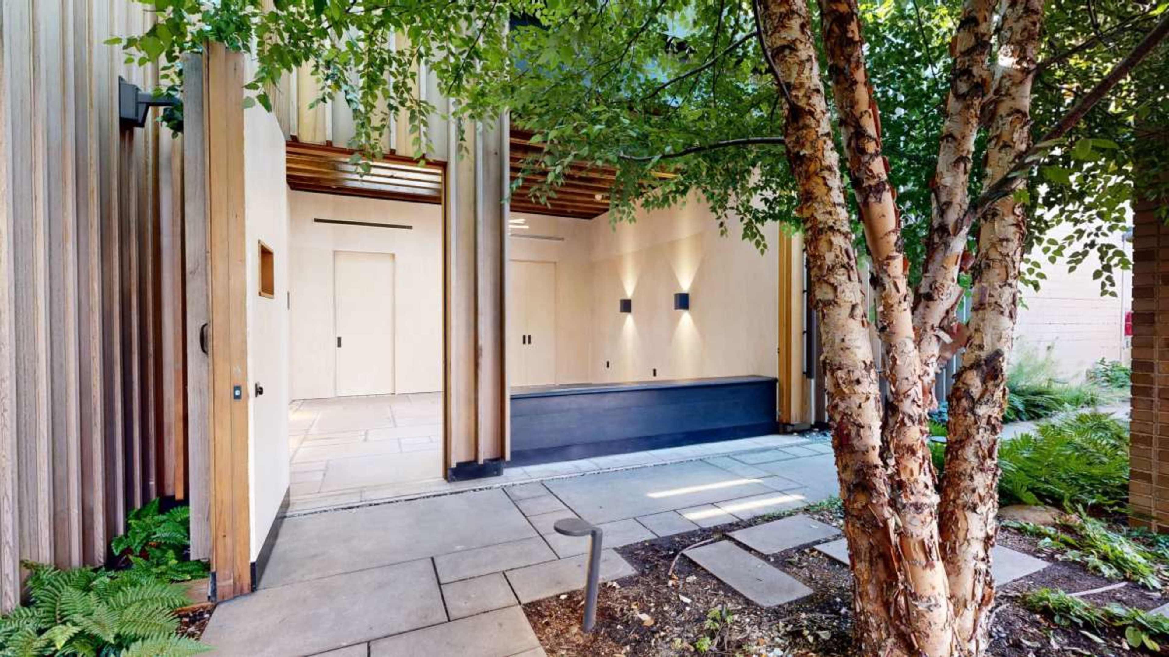 A modern entrance features wooden walls and a stone pathway, framed by greenery and a birch tree.