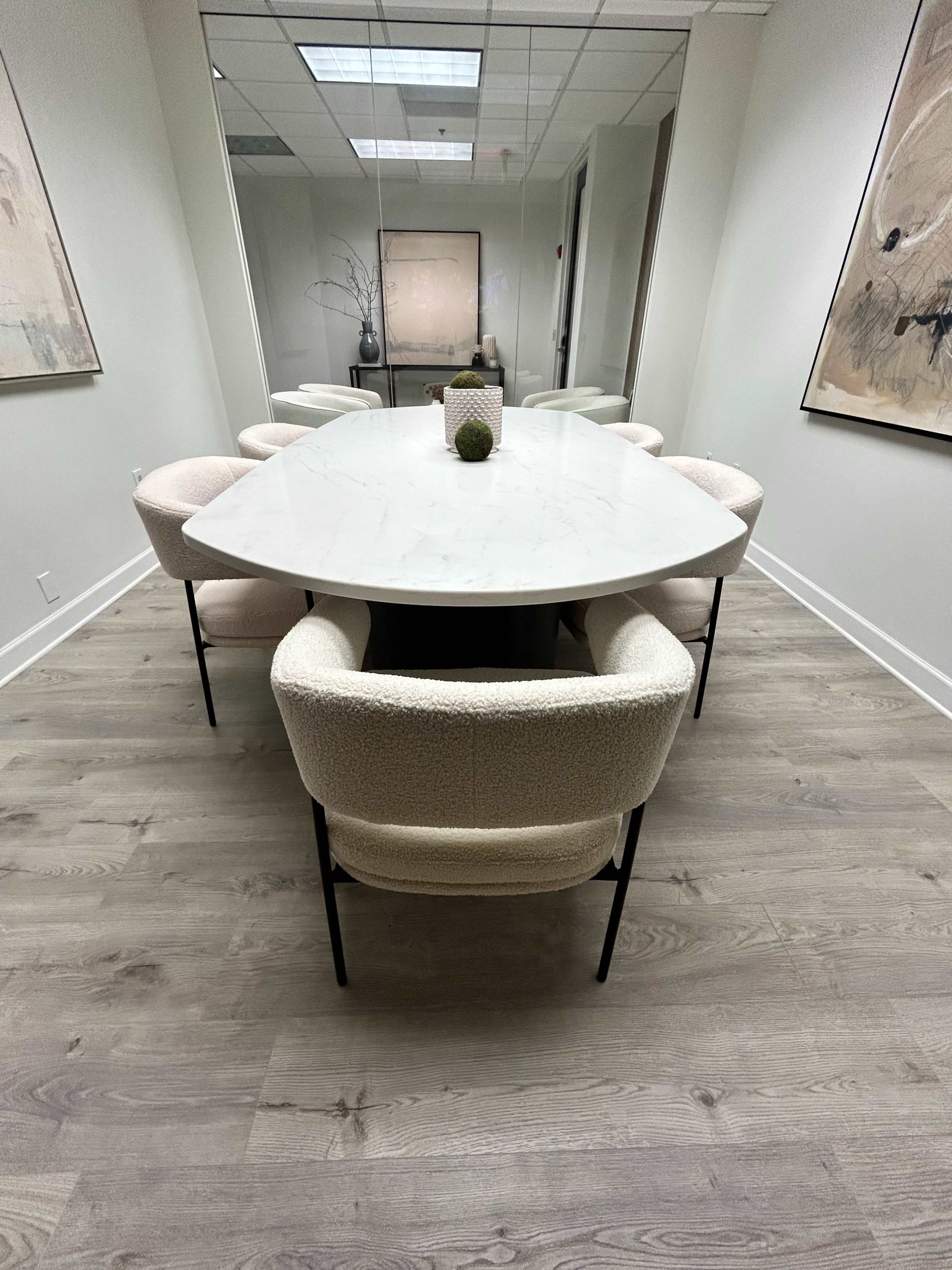 A large, oval marble table is surrounded by six upholstered chairs in a modern, minimalist conference room featuring mirrored walls and art on the sides.