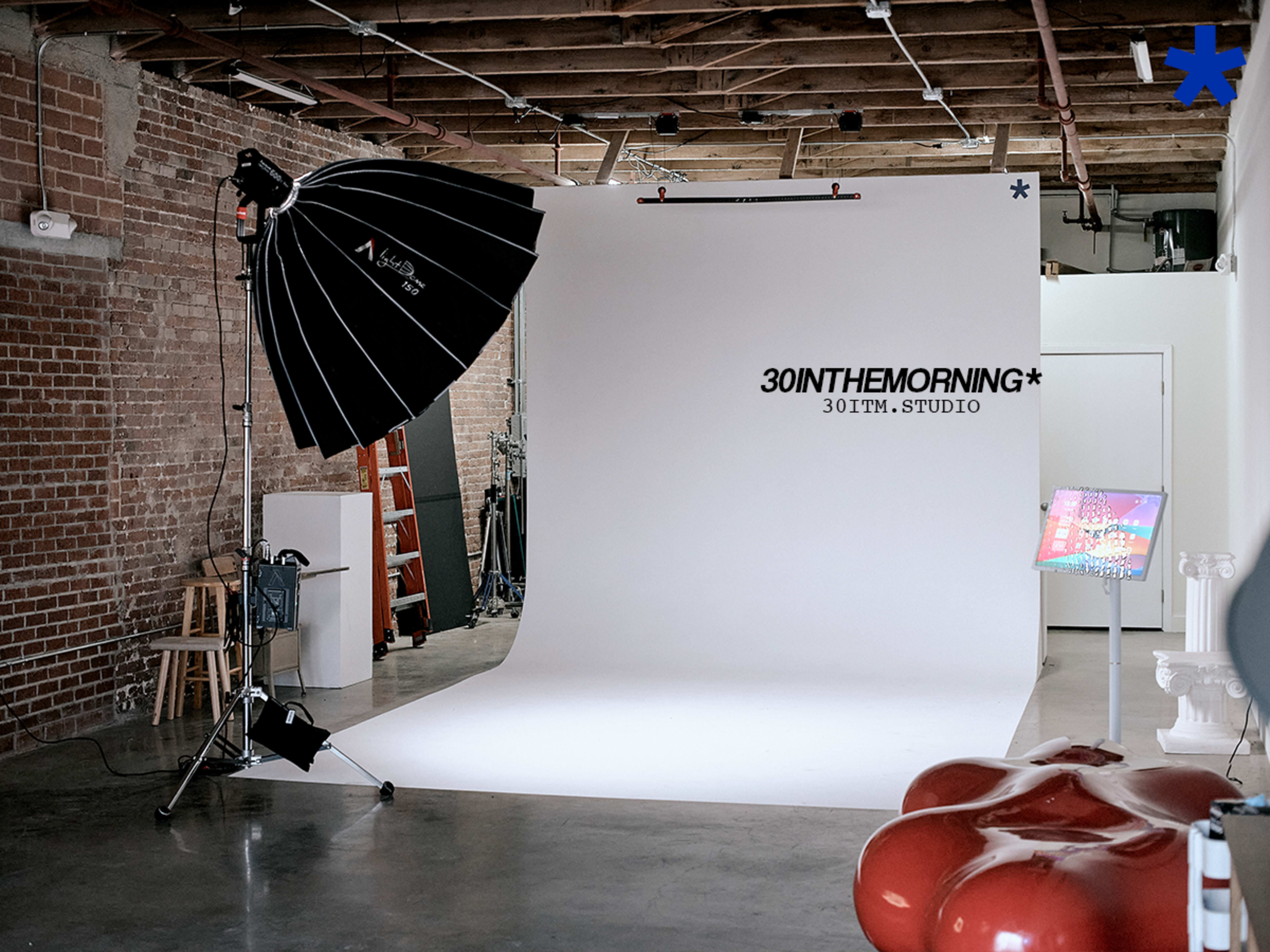 A photography studio features a large white backdrop, a softbox light, and various studio equipment in a spacious room with exposed brick walls.