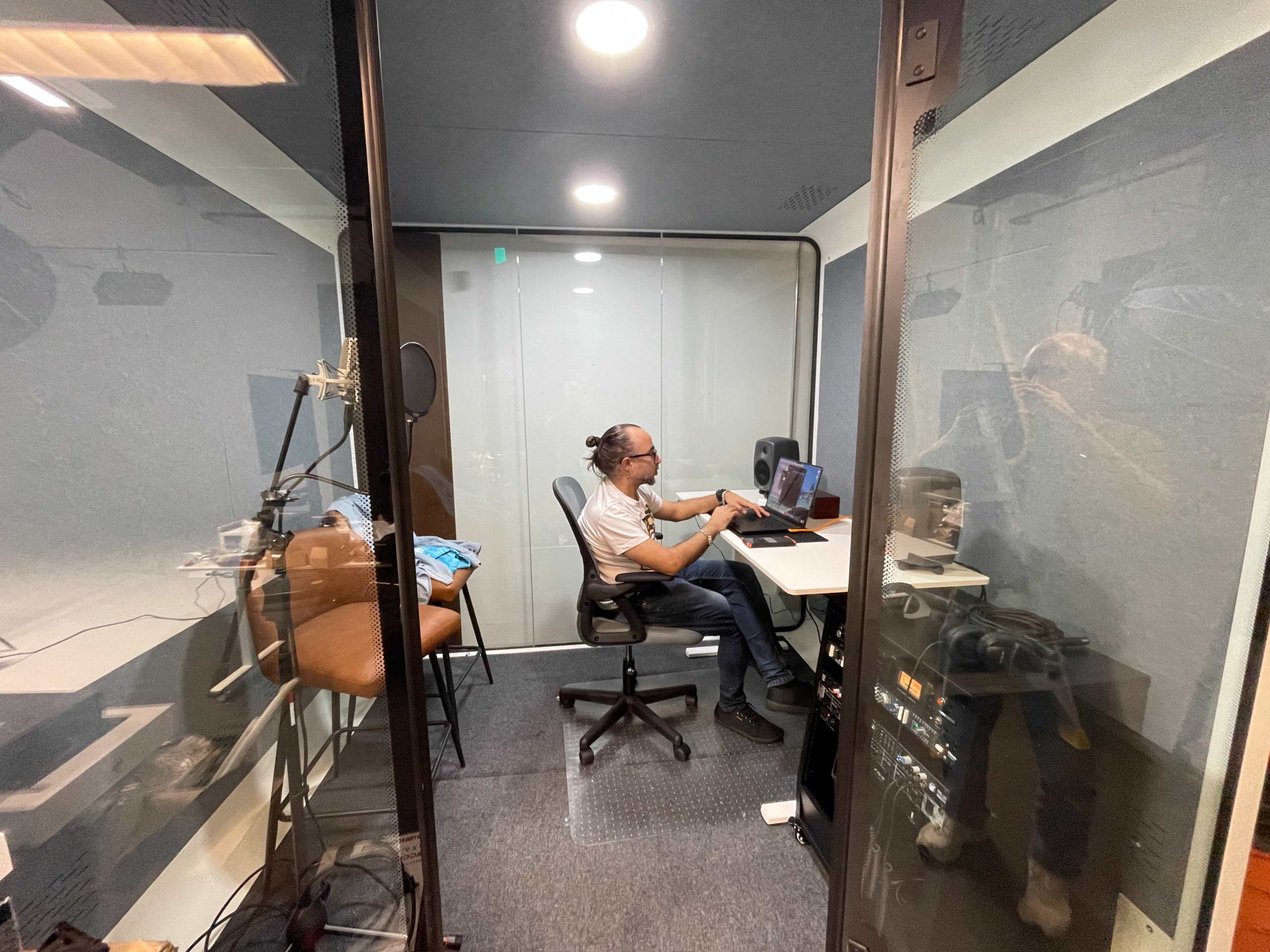 A person is working at a desk inside a soundproof recording booth equipped with audio equipment.
