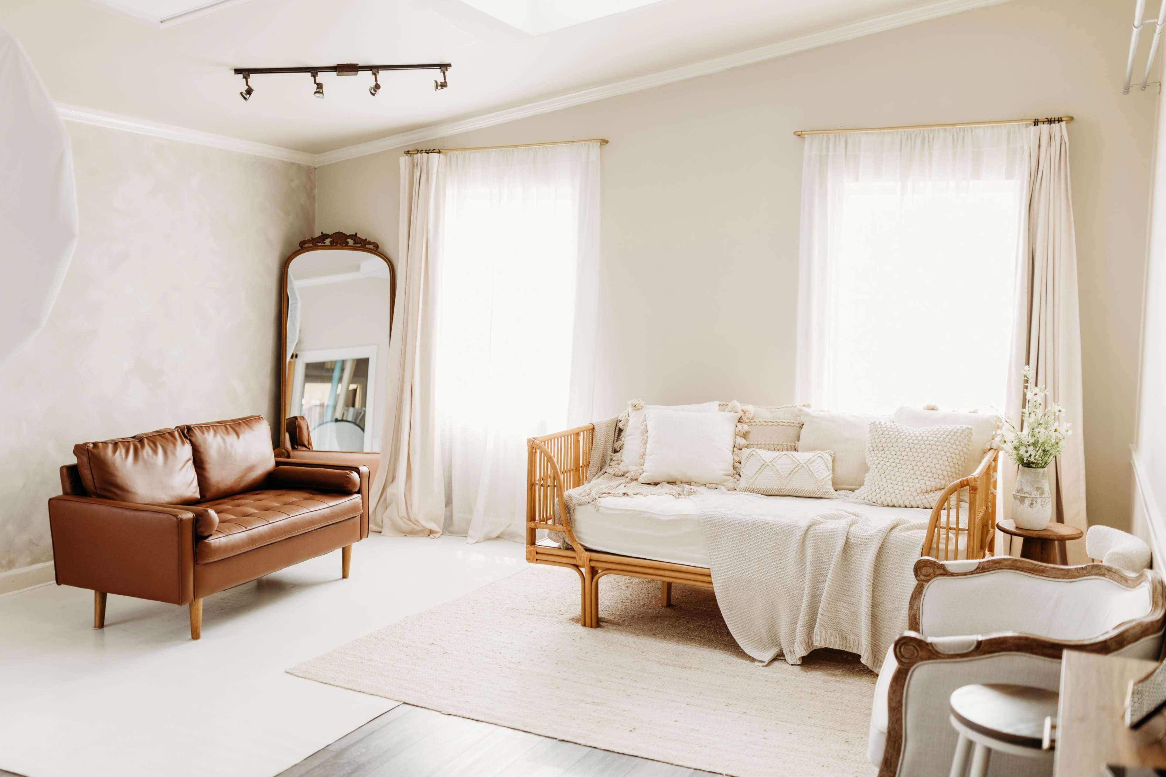 The image shows a well-lit living room featuring a brown leather sofa, a wooden daybed with cushions, and large windows draped with sheer curtains.