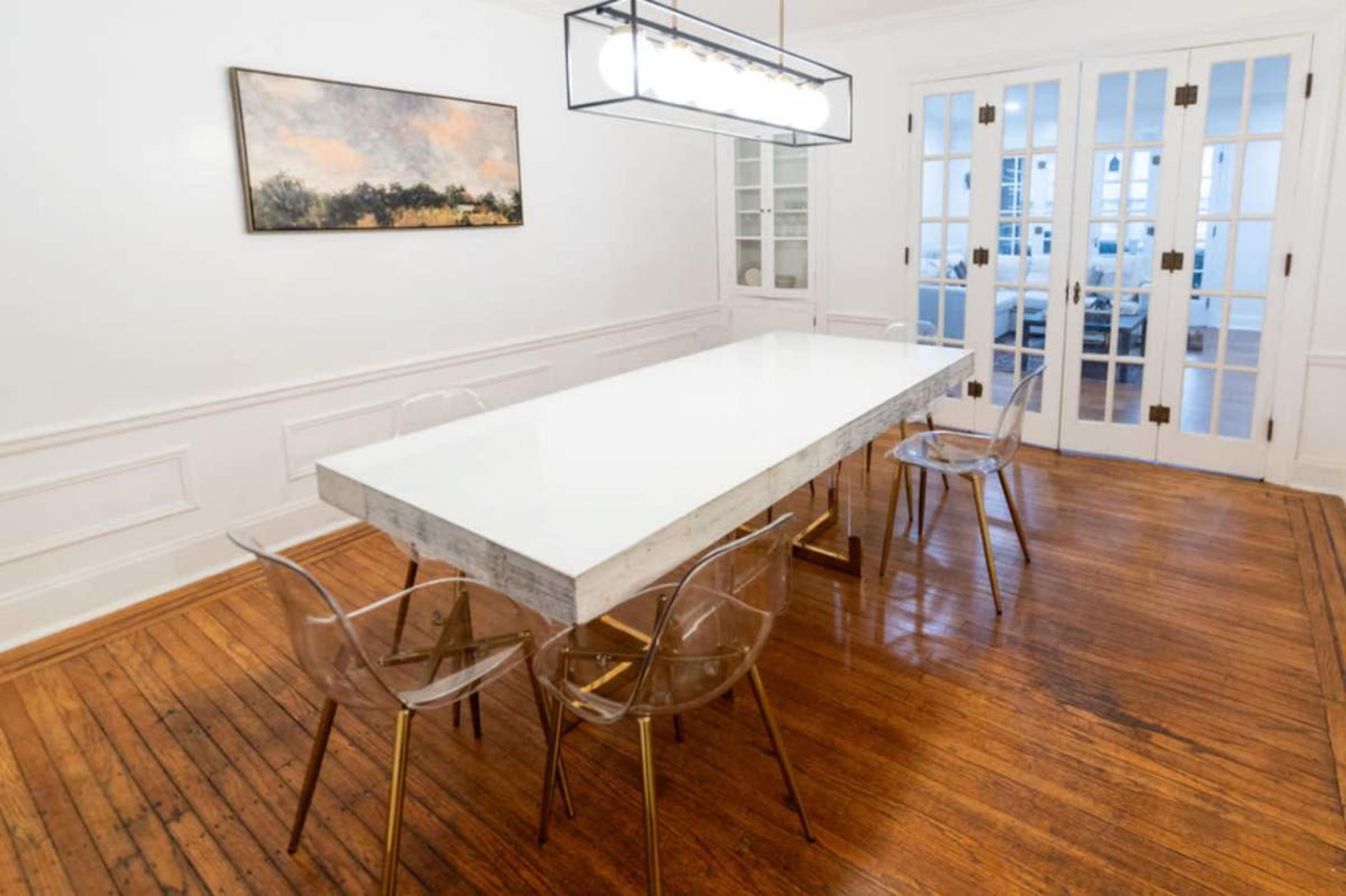 A long white dining table with clear chairs is centered in a brightly lit room featuring a wooden floor and a painting on the wall.