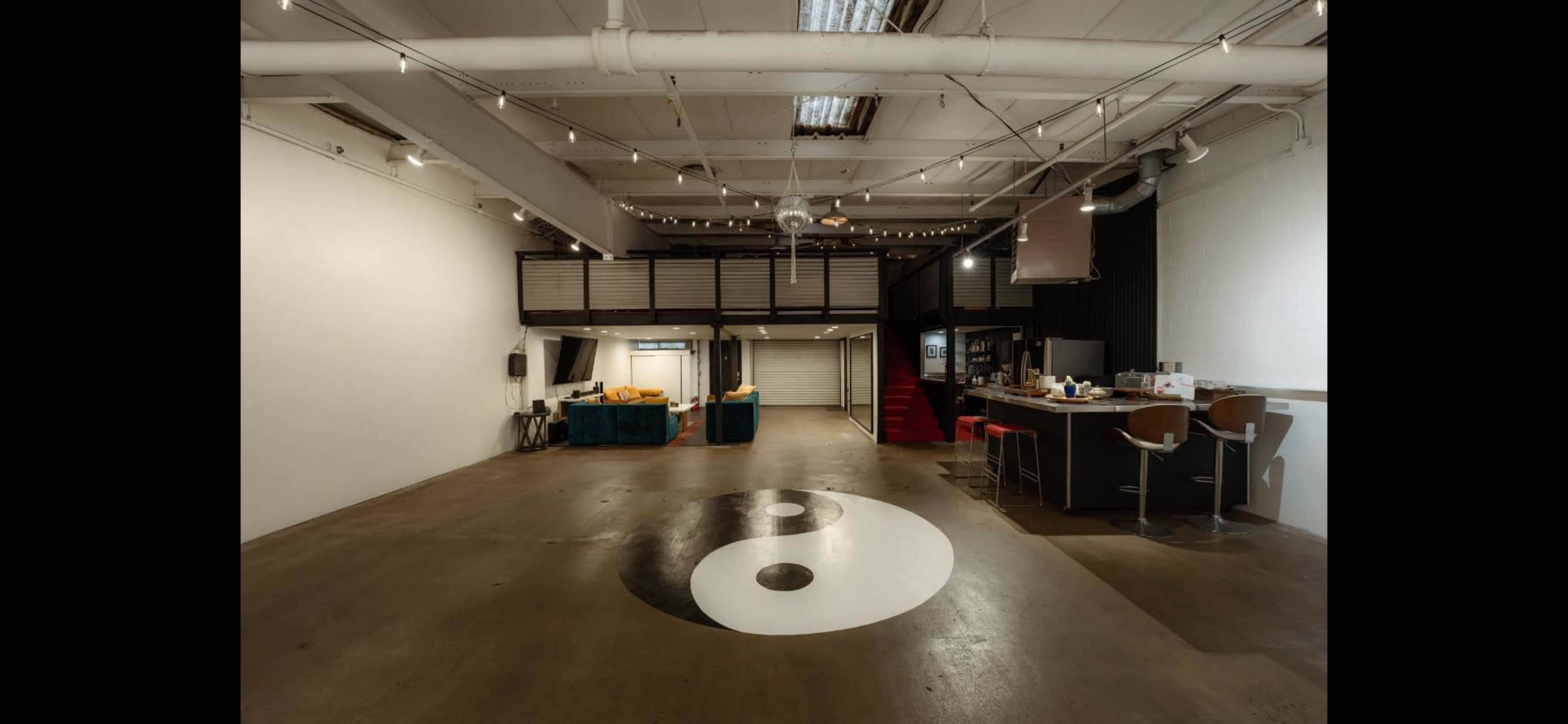 A spacious, industrial-style interior featuring a prominent yin-yang symbol on the floor, a kitchenette area, and a staircase leading to a lofted space.