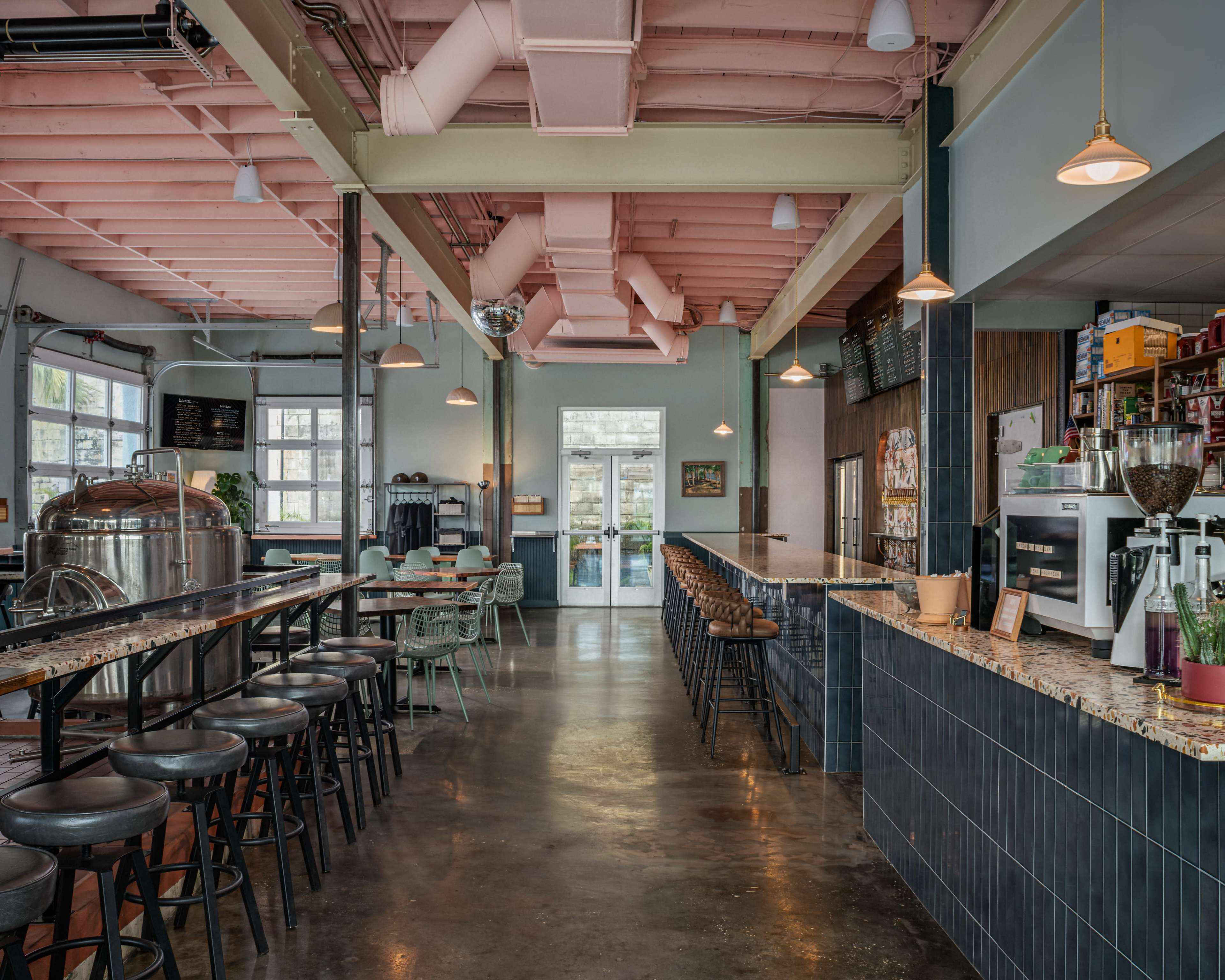 The image shows a modern café interior with a long bar counter, seating area, and large windows allowing natural light to enter.