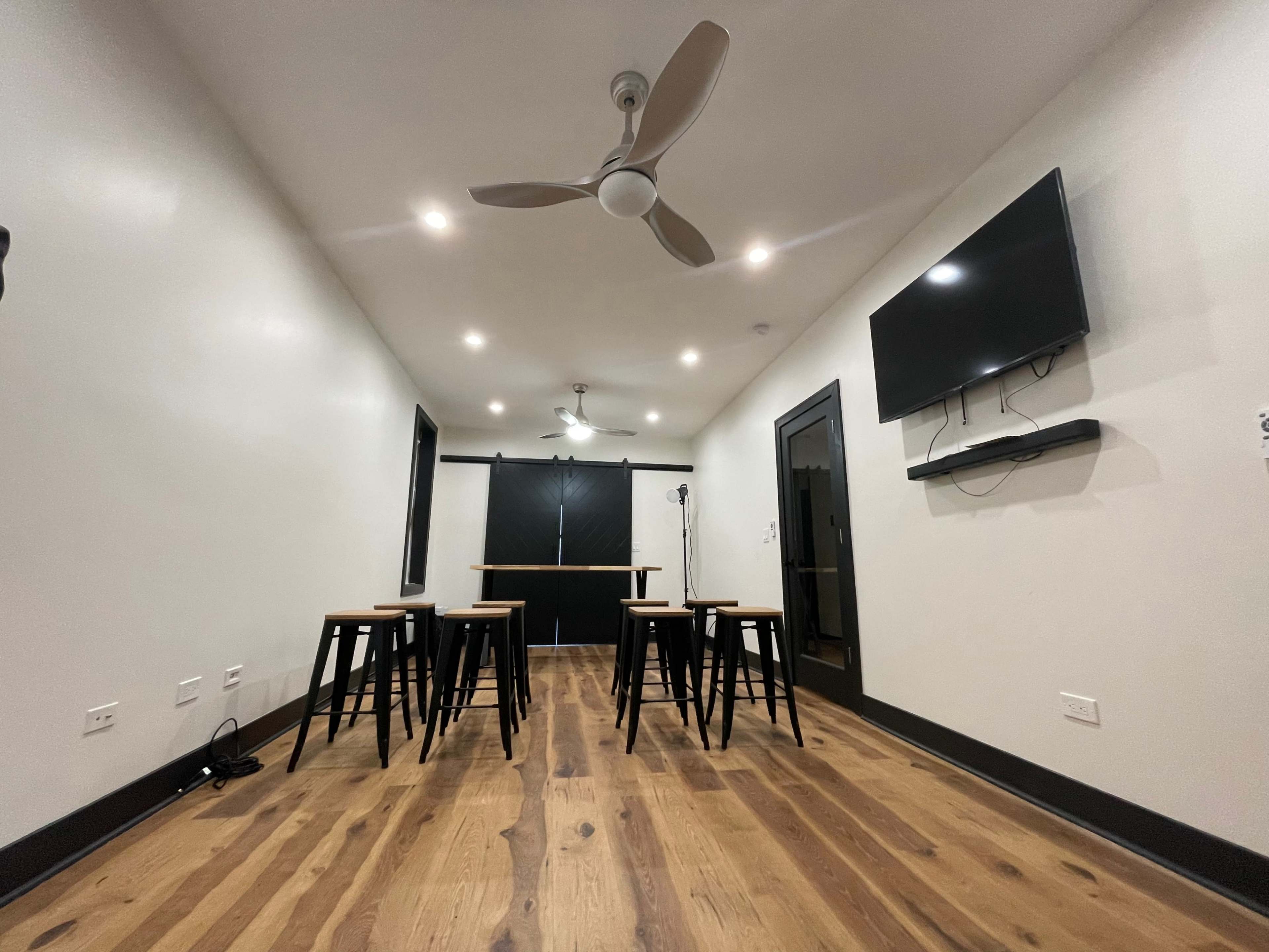 The image shows a modern room with a wooden floor, a table in the center, several black stools, a wall-mounted TV, and a ceiling fan.