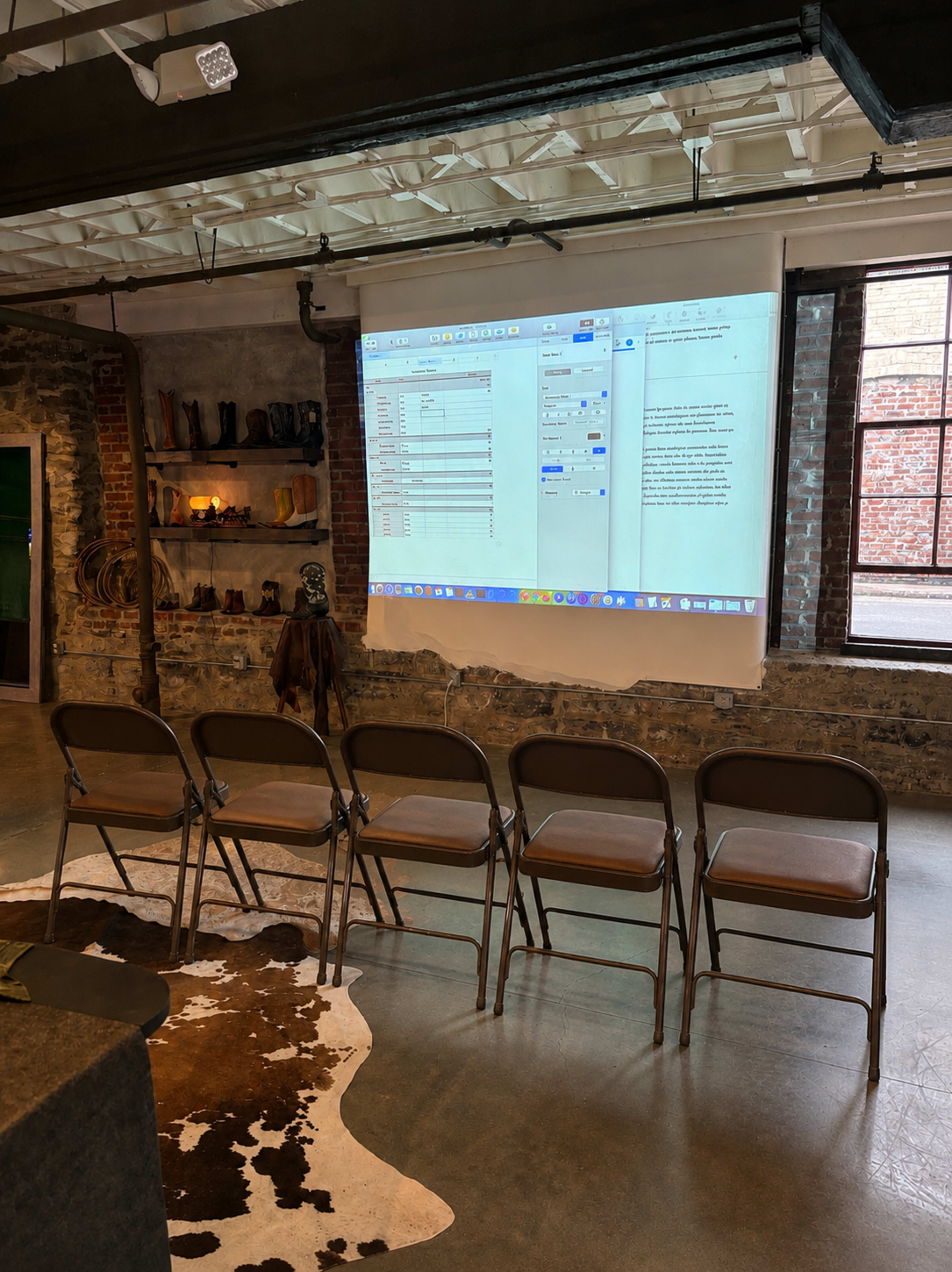 A row of metal folding chairs is arranged facing a large screen displaying a spreadsheet and a document in a brick and mortar room with rustic decor.