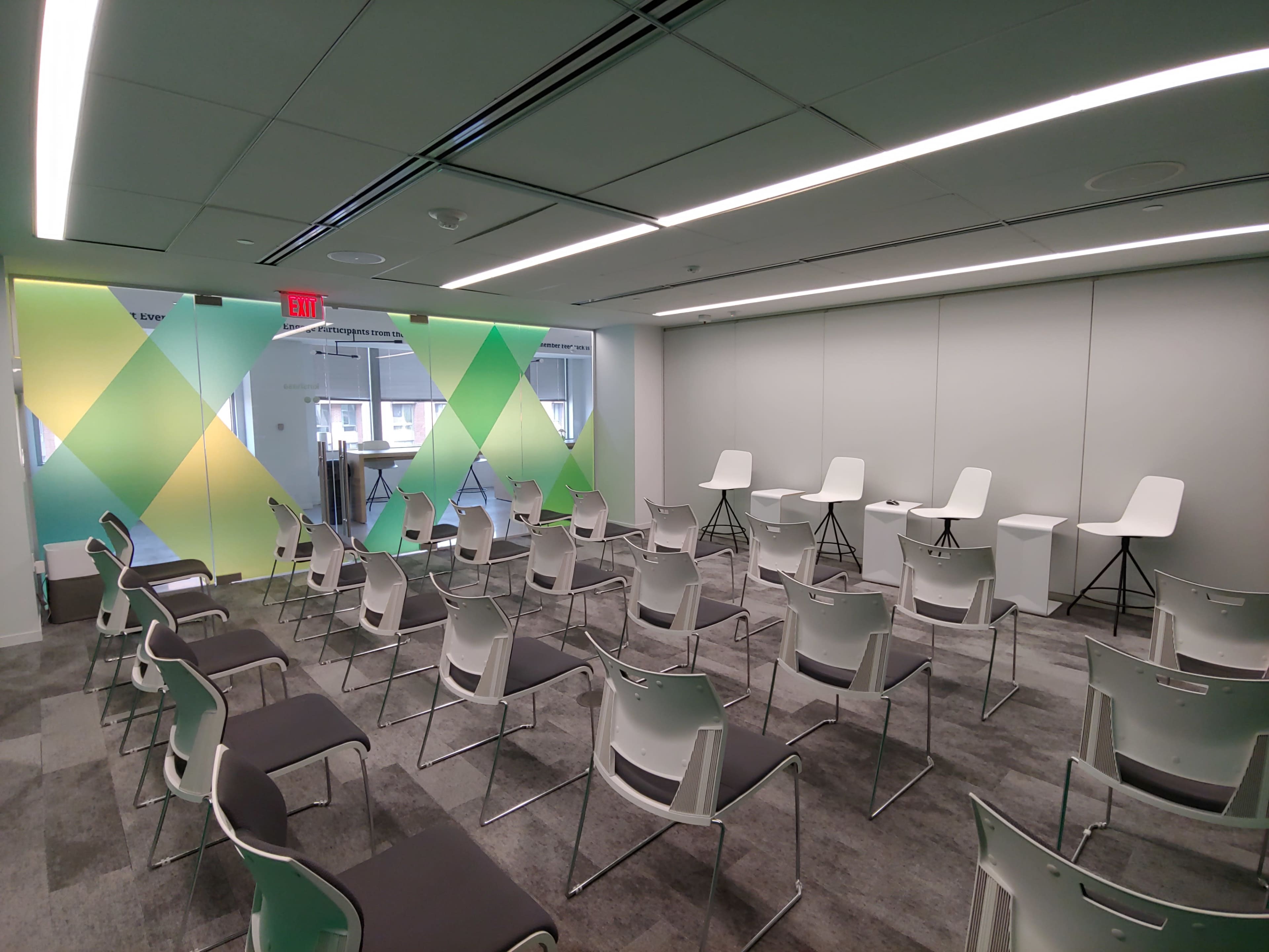 A meeting room with neatly arranged chairs facing a small stage area, featuring colorful wall graphics and large windows.