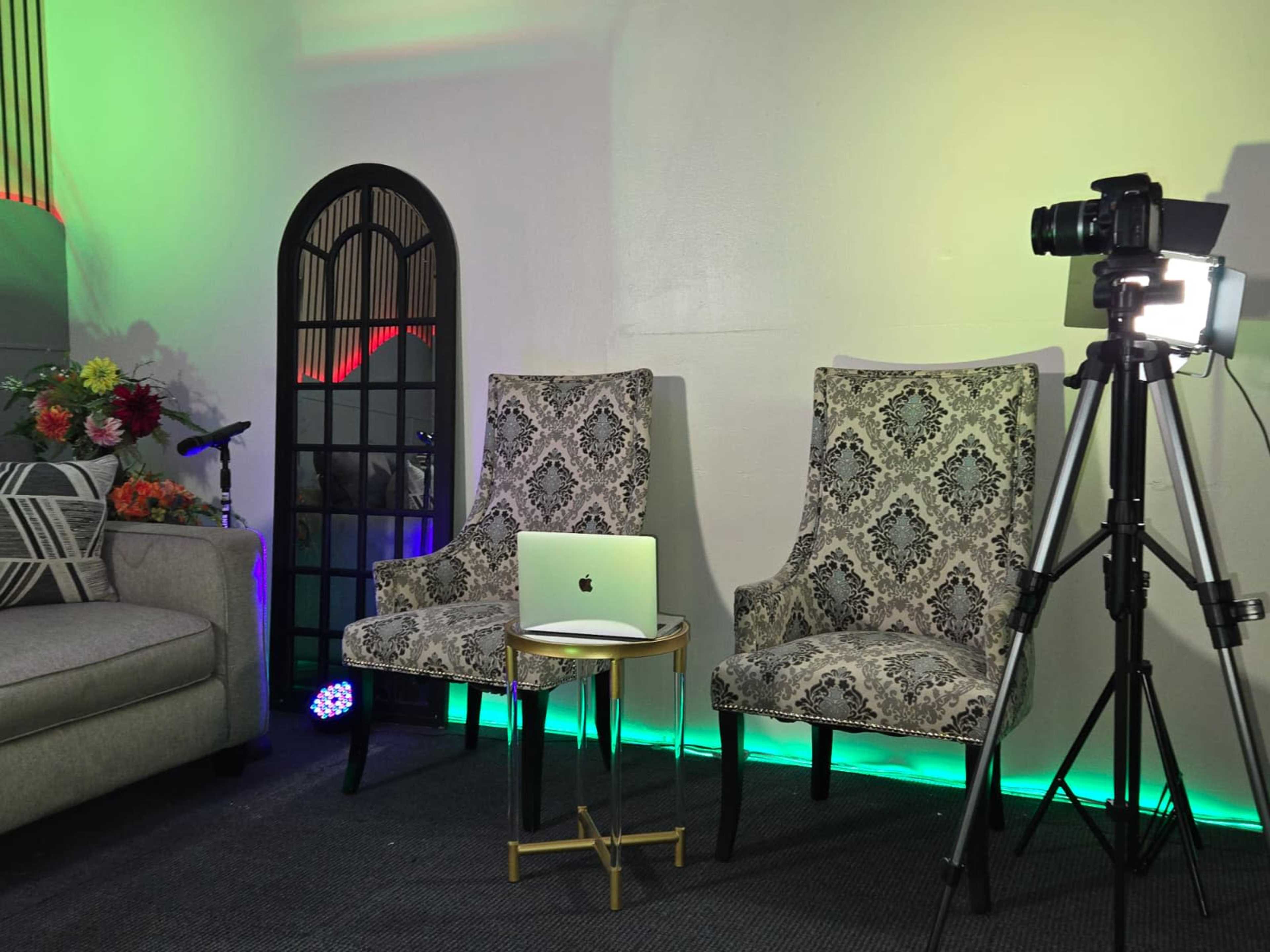 The image shows a well-lit indoor setup featuring two patterned chairs, a small table with a laptop, and a camera mounted on a tripod, with decorative elements in the background.