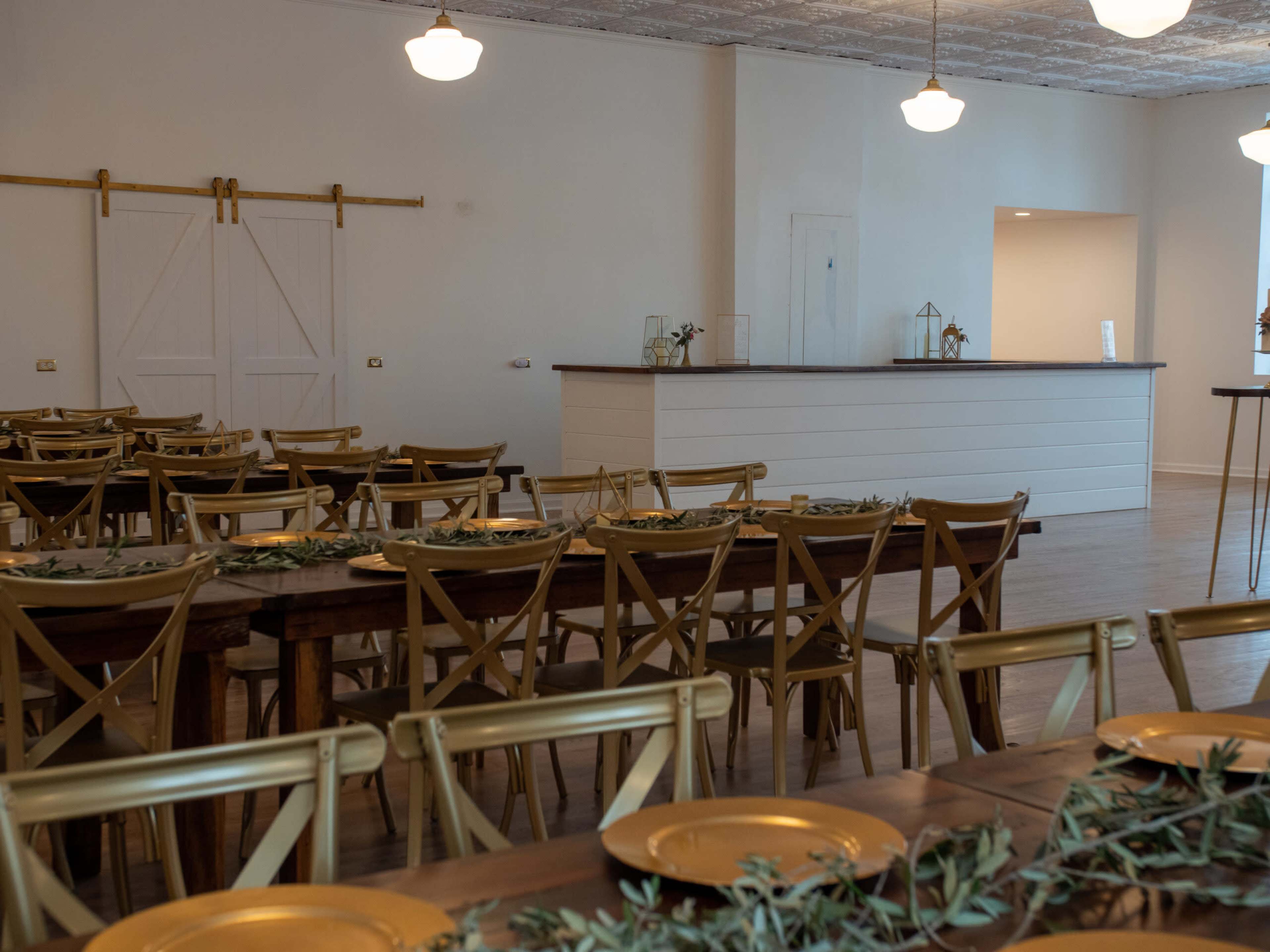 A spacious event room features wooden tables set with gold-rimmed plates and rows of chairs facing a simple front counter.