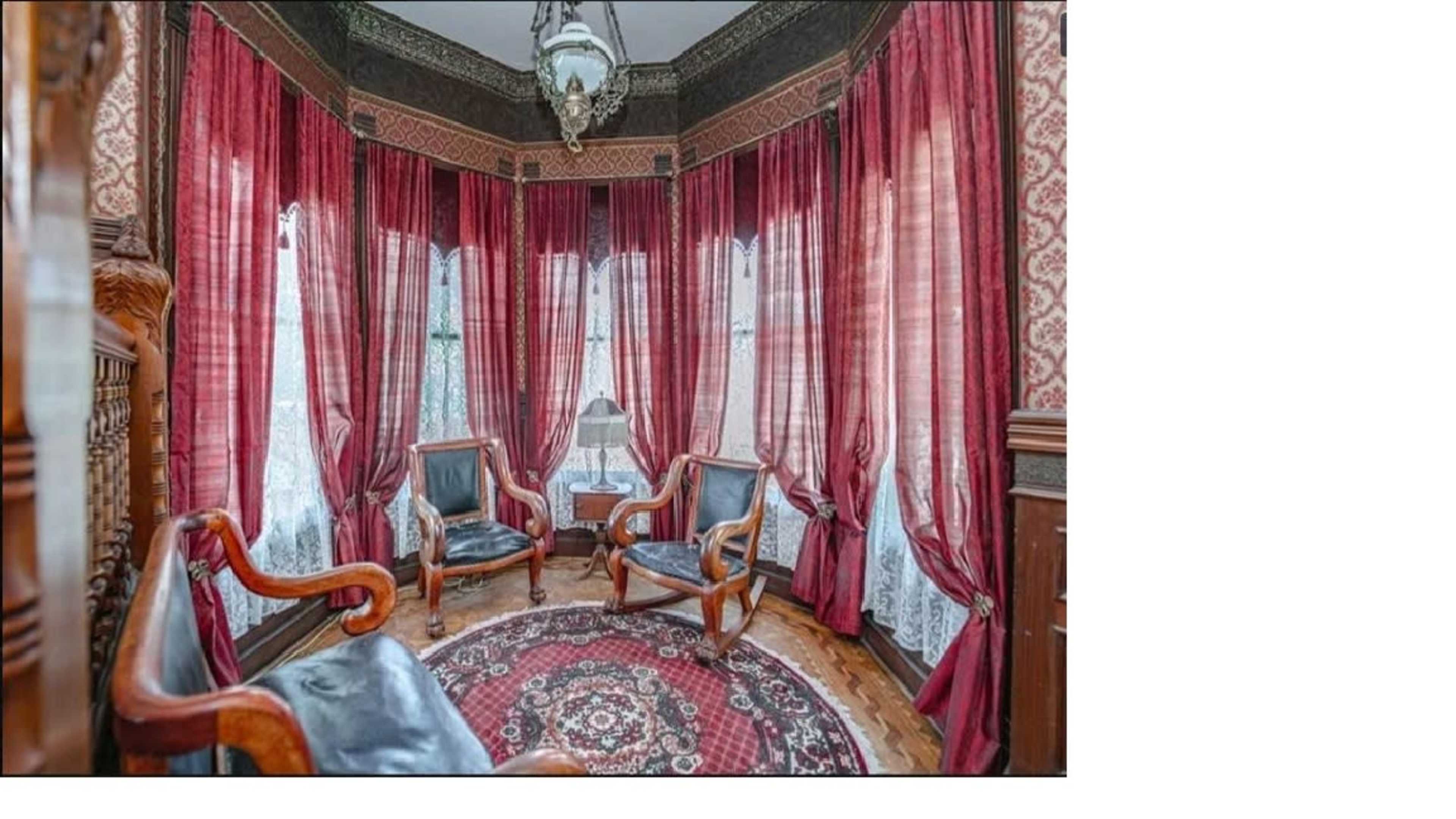 A room with large windows draped in red curtains, featuring three leather chairs arranged around a circular rug.