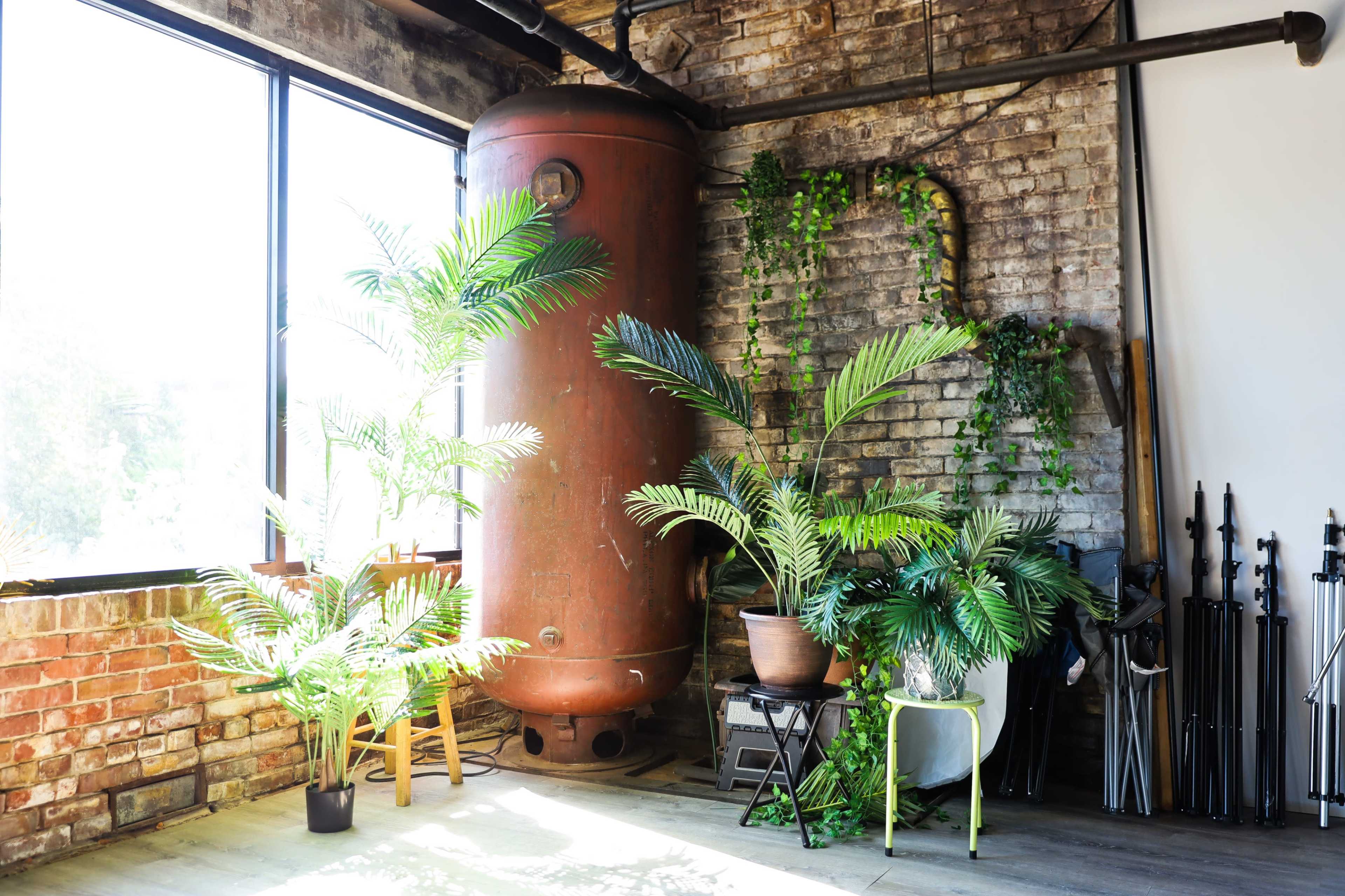 A large red water tank alongside several potted plants in a bright room with exposed brick walls and large windows.