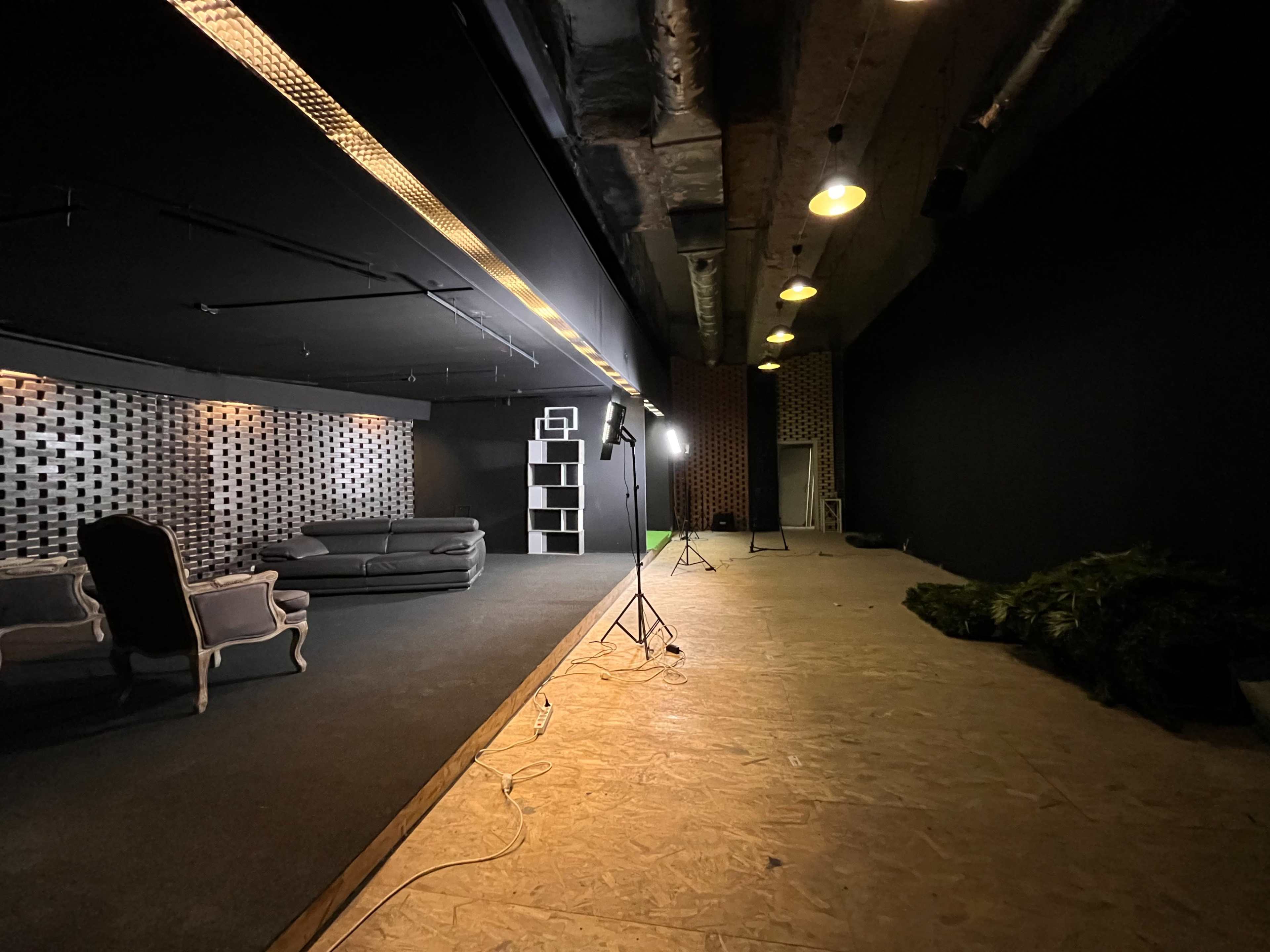 The image shows a spacious, dimly lit room featuring a gray couch, a vintage chair, a backdrop of patterned wall panels, and a light stand on a wooden floor.