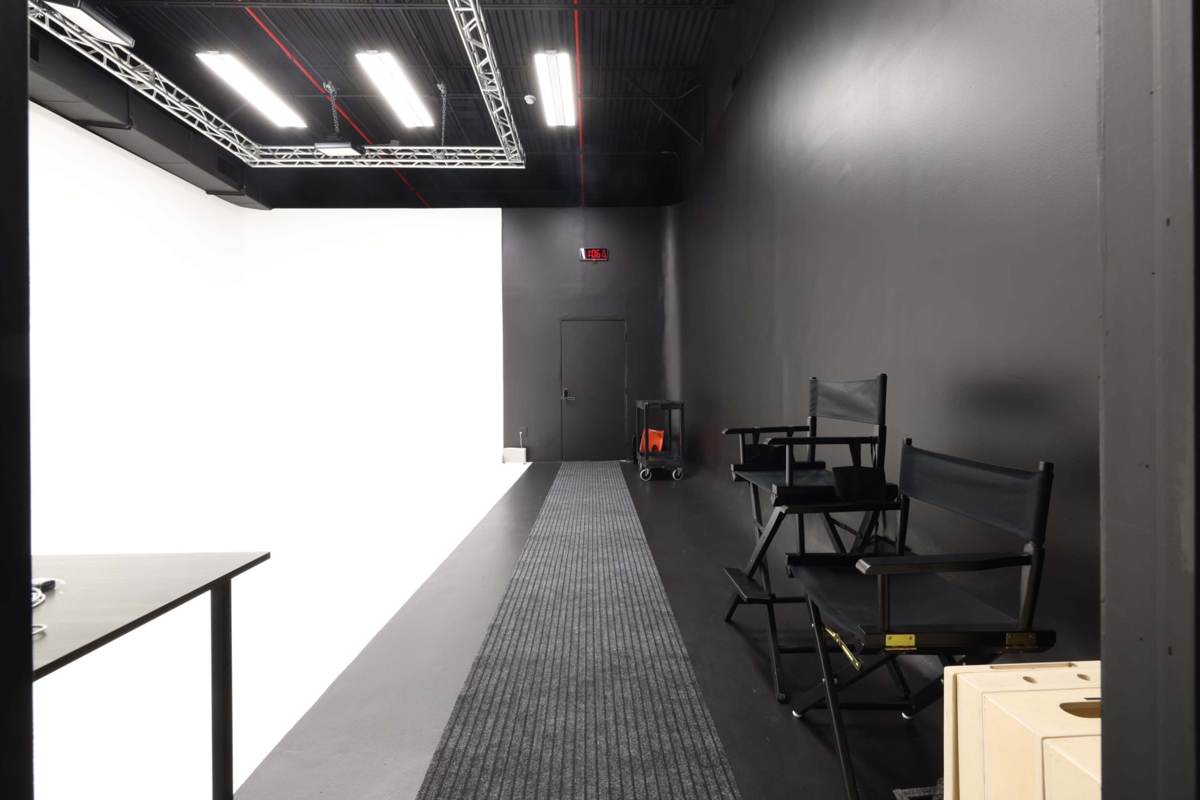 The image shows a narrow hallway leading to a large white studio space, flanked by black walls and featuring a few director's chairs and a table.