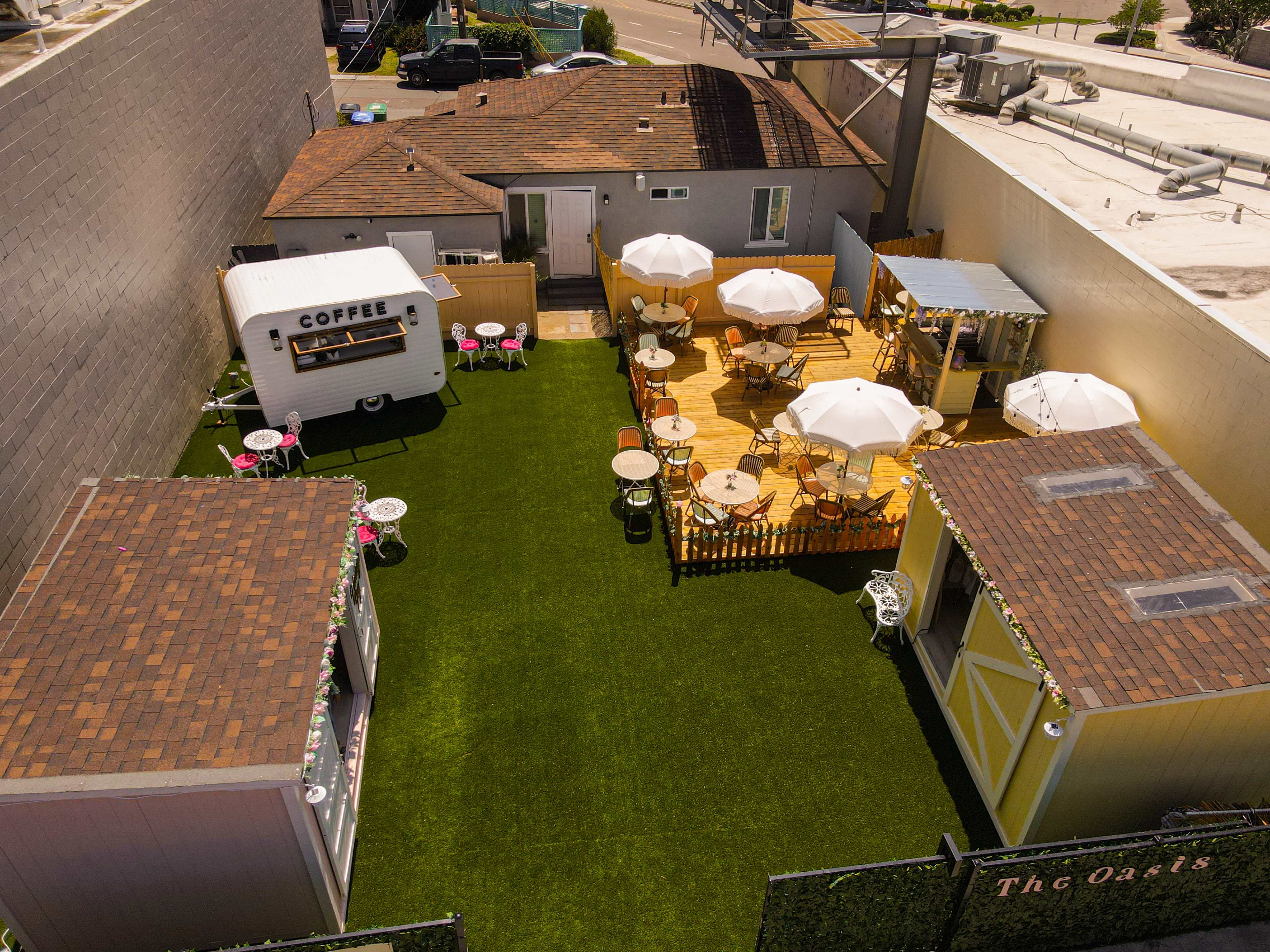 A small outdoor café area with a coffee trailer, shaded tables, and a grassy space surrounded by buildings.