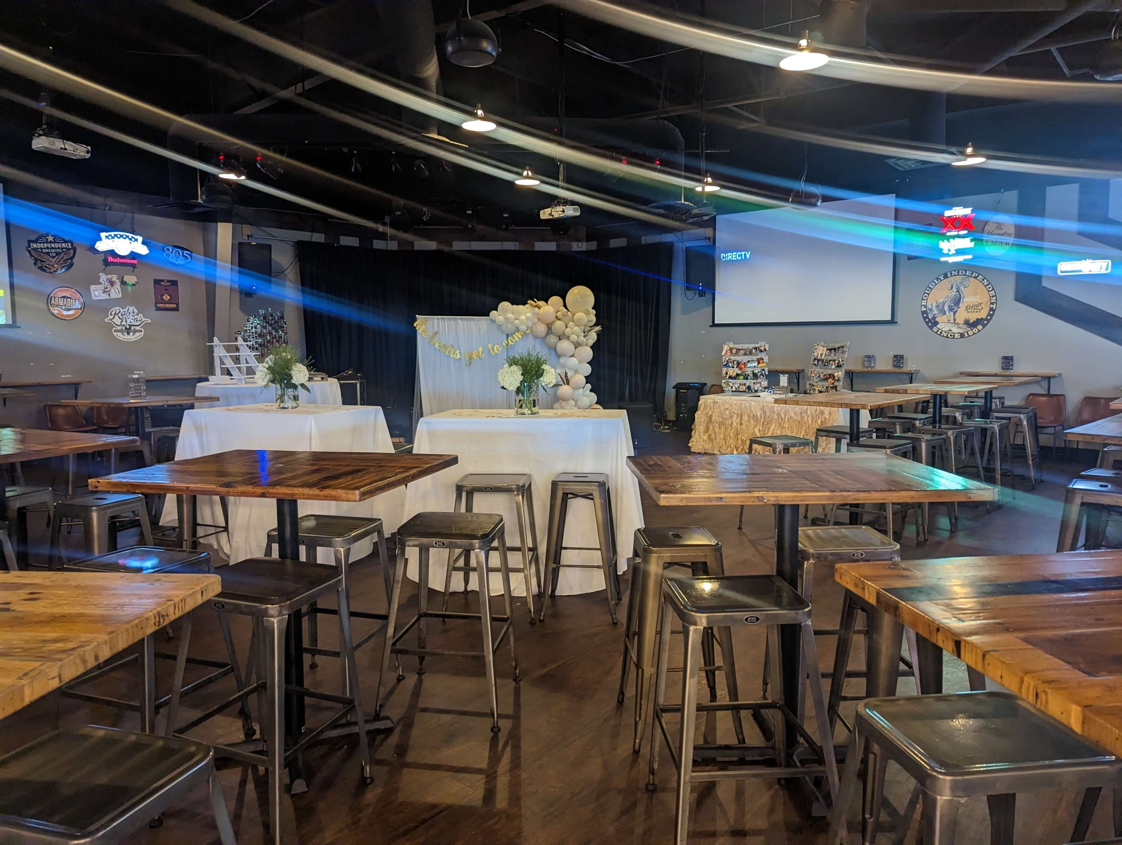 A spacious event venue with several wooden tables and chairs, featuring a decorated stage area and a display table in the background.