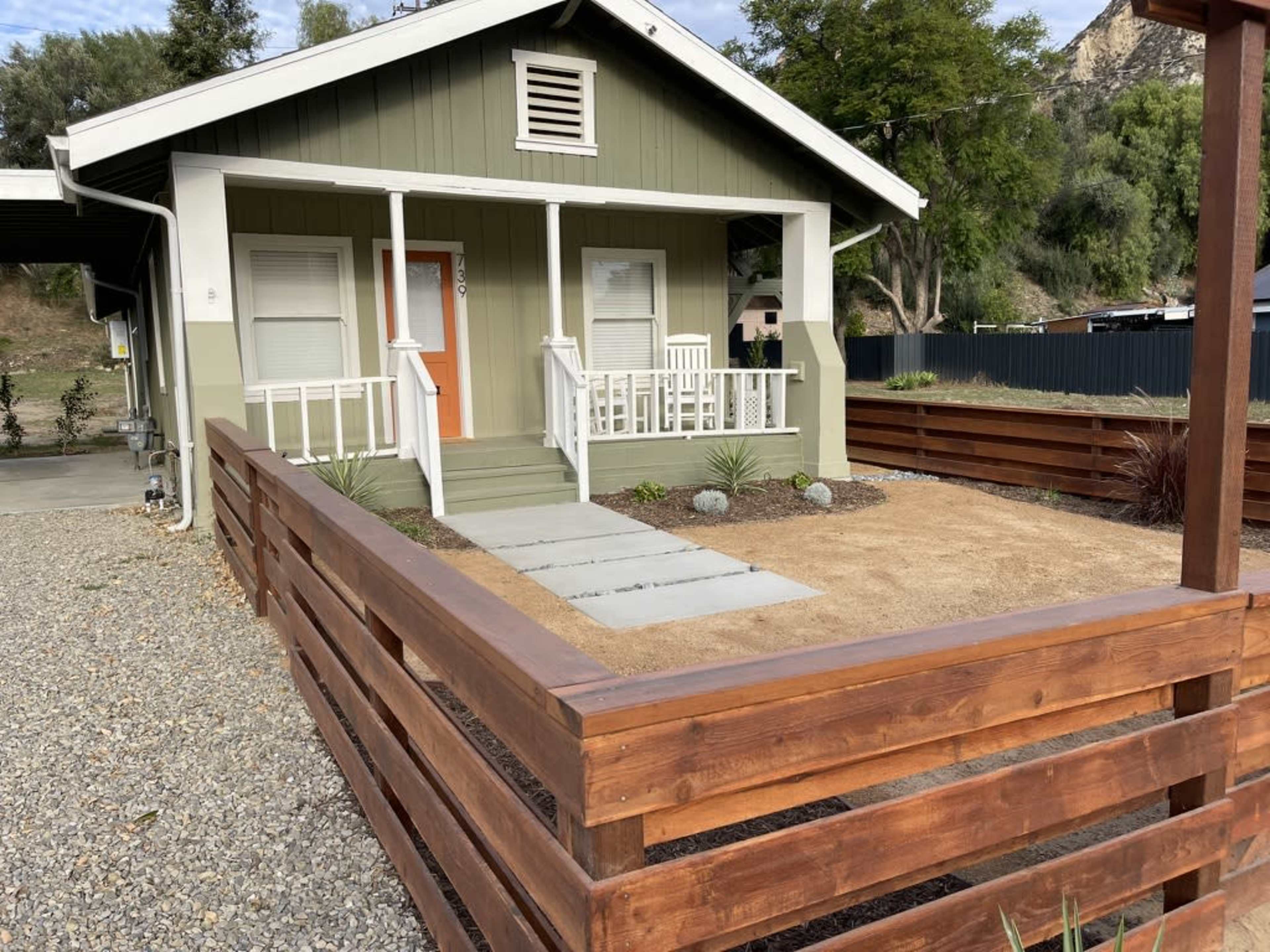A small house with a green exterior, a front porch, and a fenced yard featuring gravel and sparse landscaping.