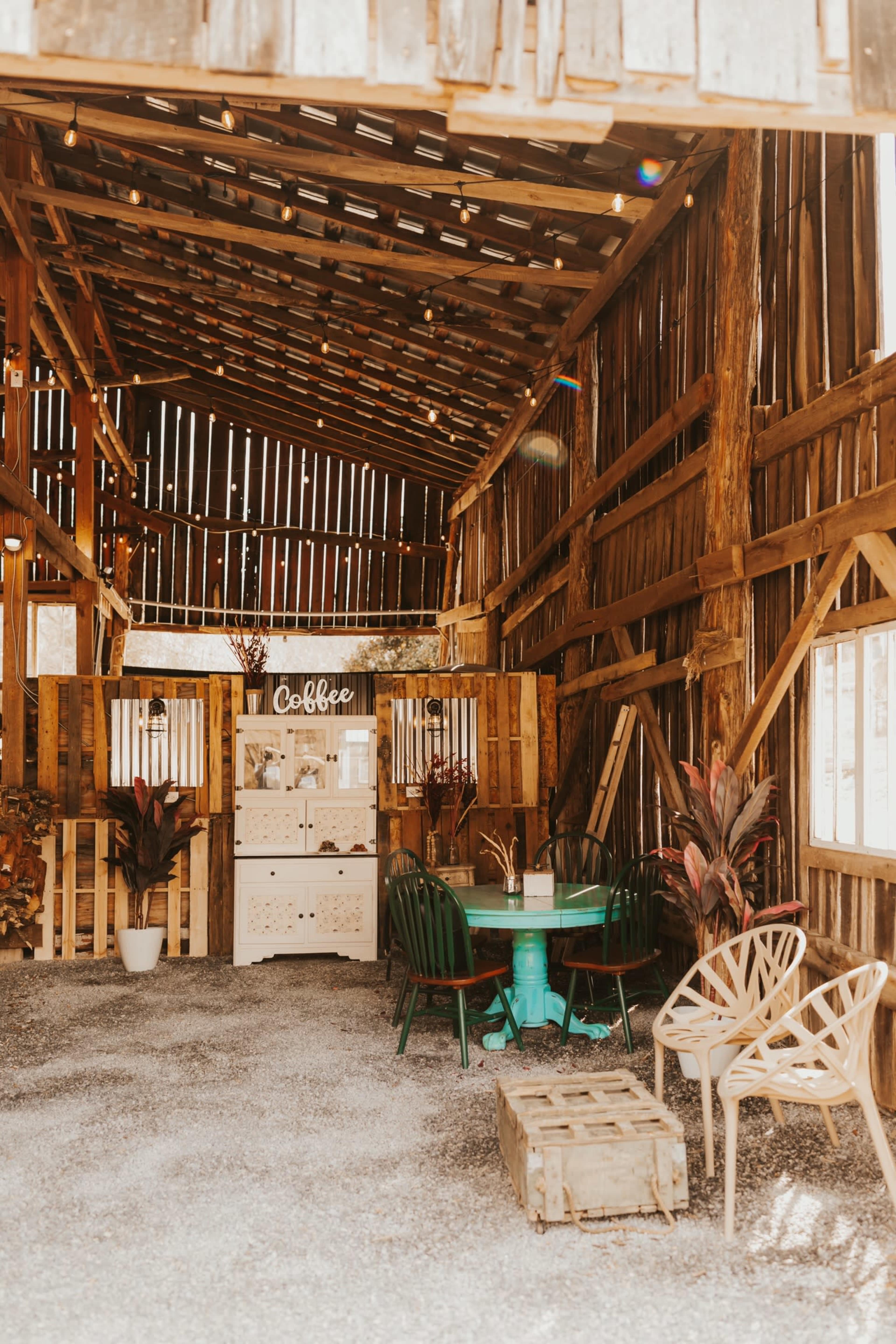 The image shows a rustic, wooden barn interior with a coffee station and seating area featuring a turquoise table and various chairs.