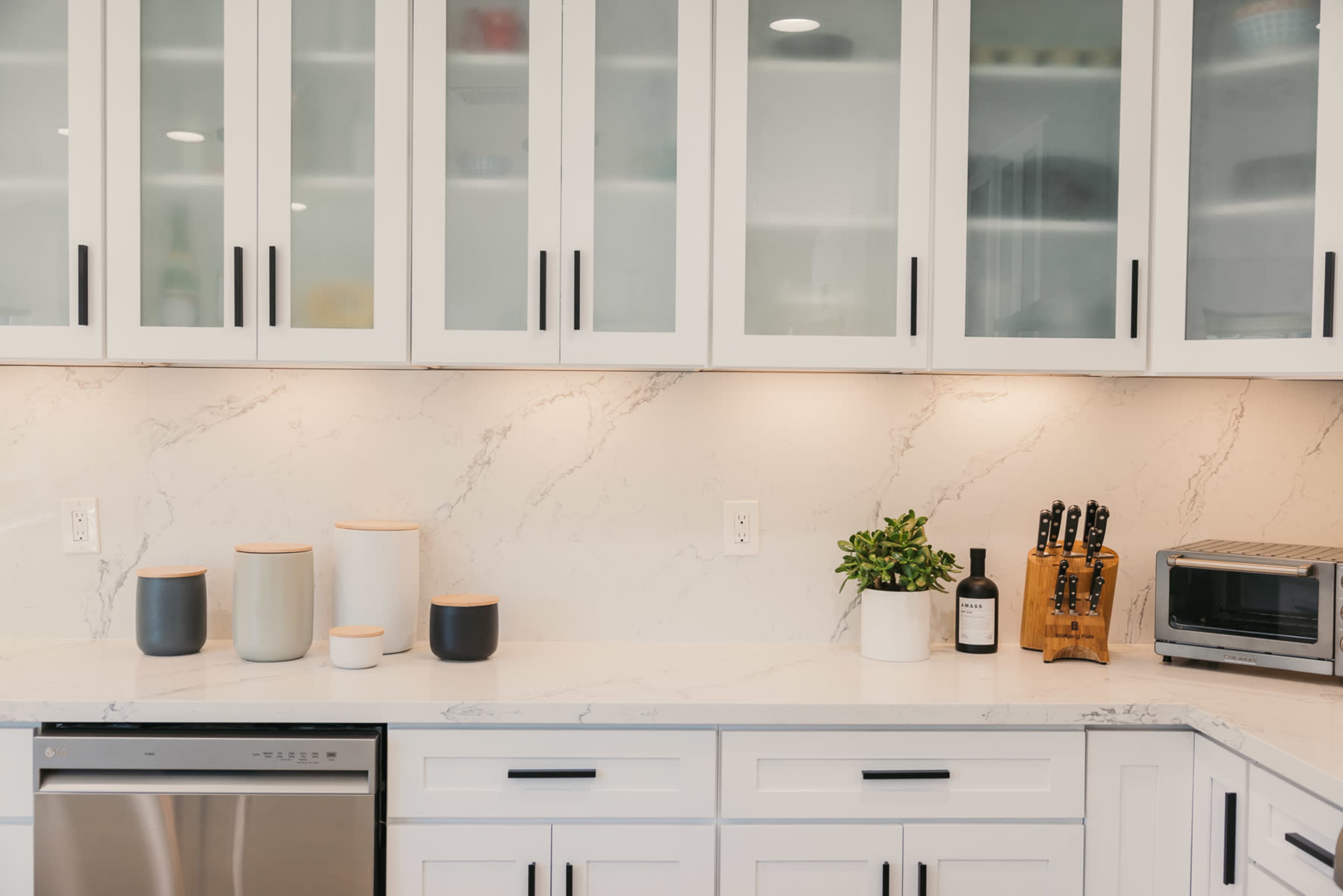 A modern kitchen features white cabinets with glass fronts, a marble countertop, various containers, a potted plant, a wooden knife block, and a microwave.