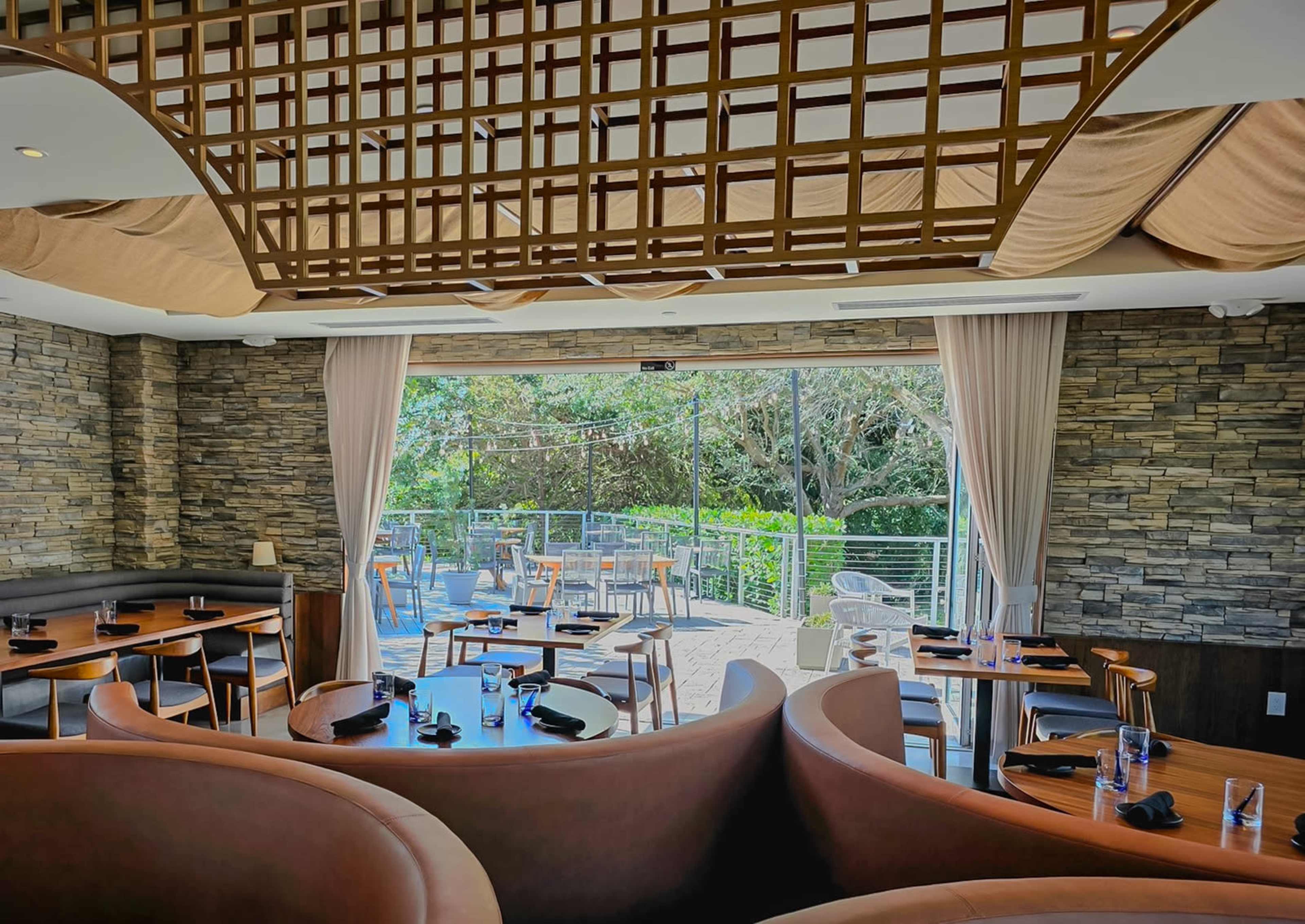 The image shows a modern restaurant interior with curved seating, stone walls, and large windows overlooking an outdoor dining area.