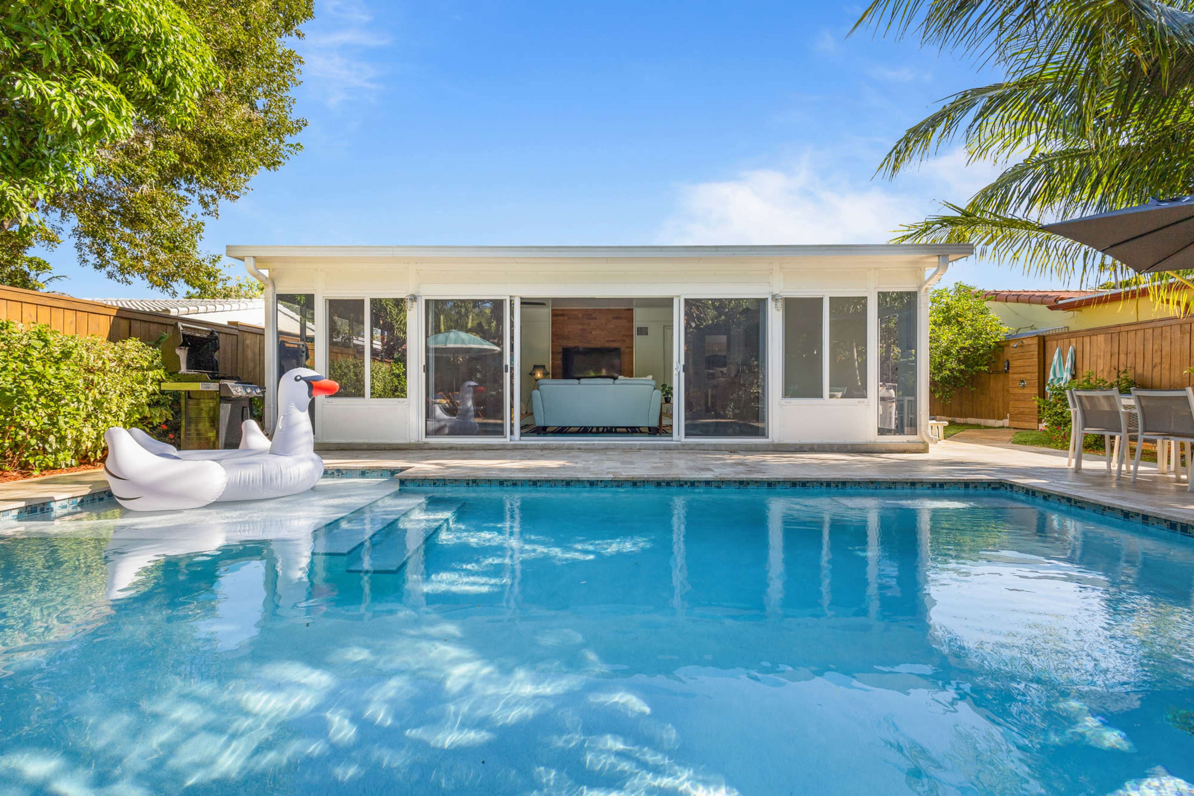 A swimming pool with a swan-shaped float is located in front of a modern house surrounded by palm trees.