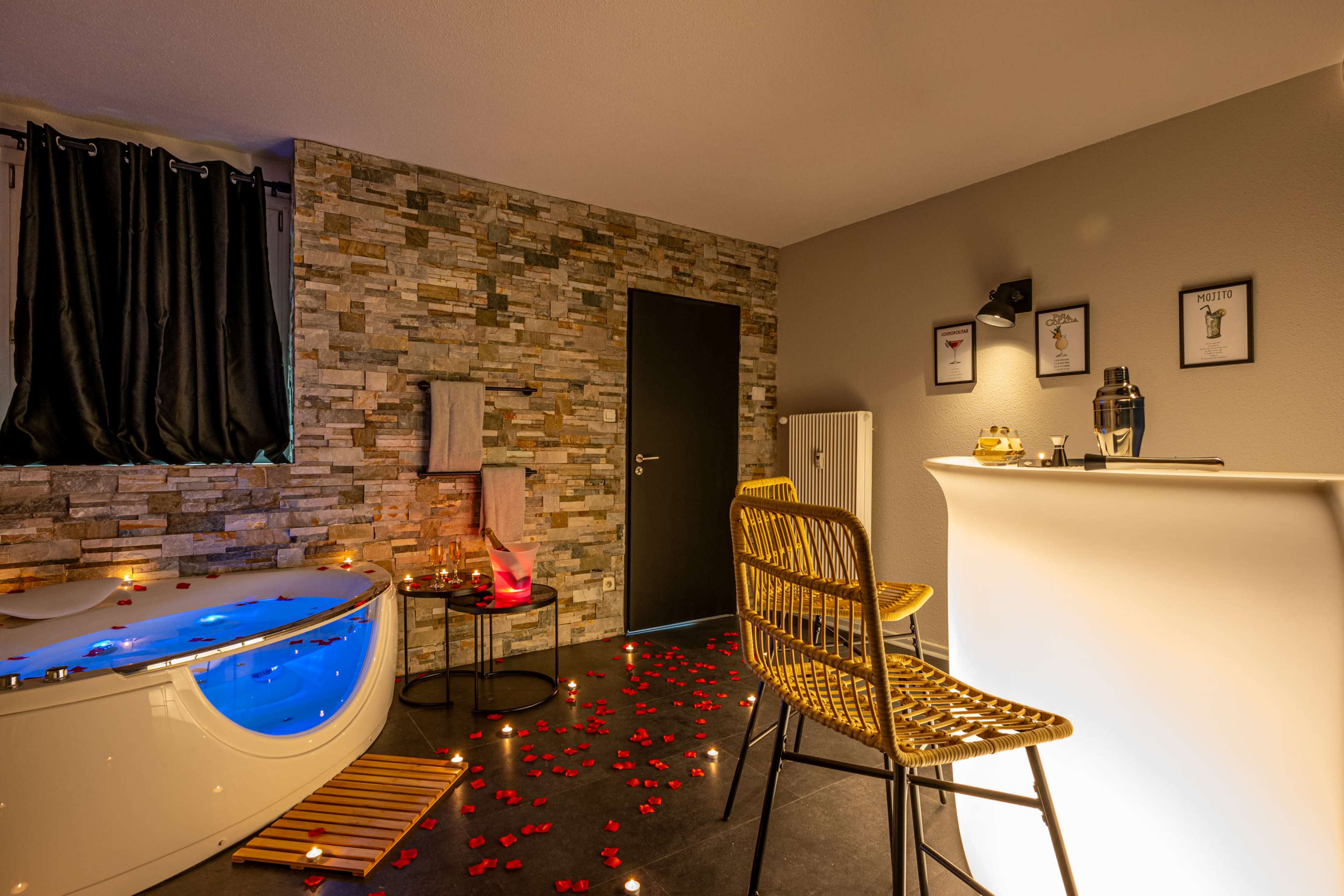 A modern bathroom with a freestanding tub, decorative stone walls, and a bar area featuring a chair and a small table adorned with rose petals and candles.