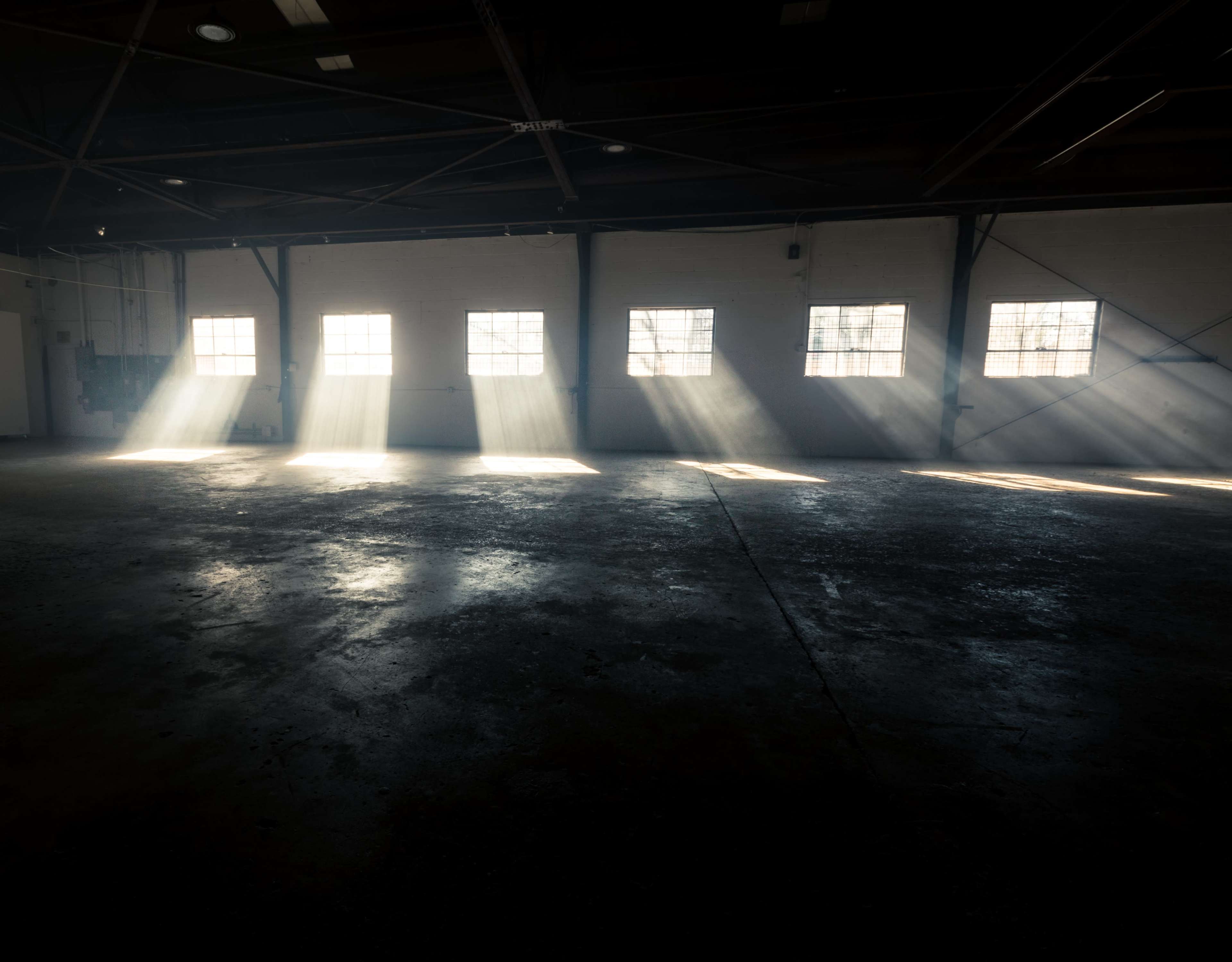 Sunlight streams through the windows of an empty industrial space, casting beams on the concrete floor.
