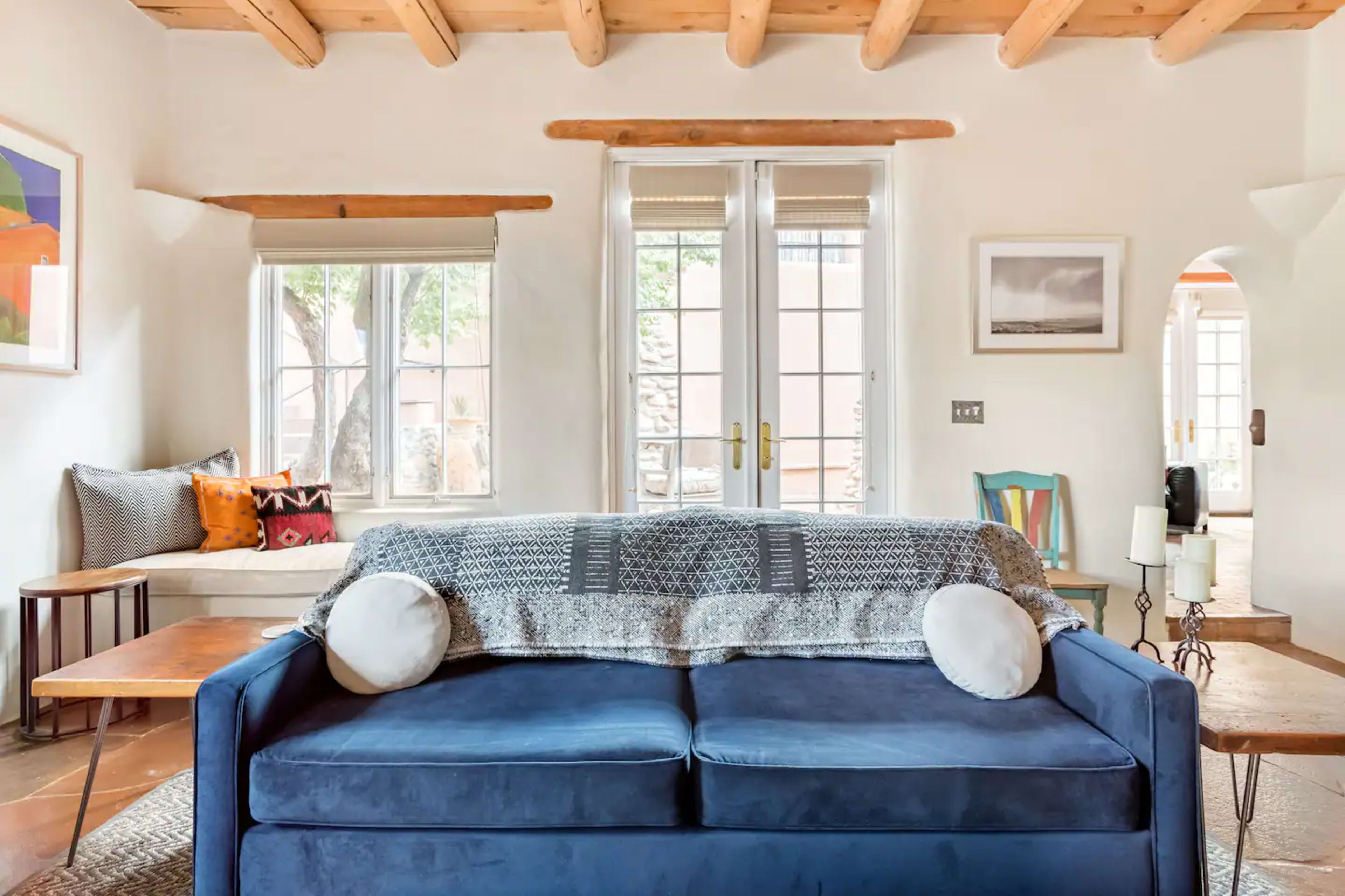 A blue sofa is centered in a brightly lit living room with large windows and wooden beams.
