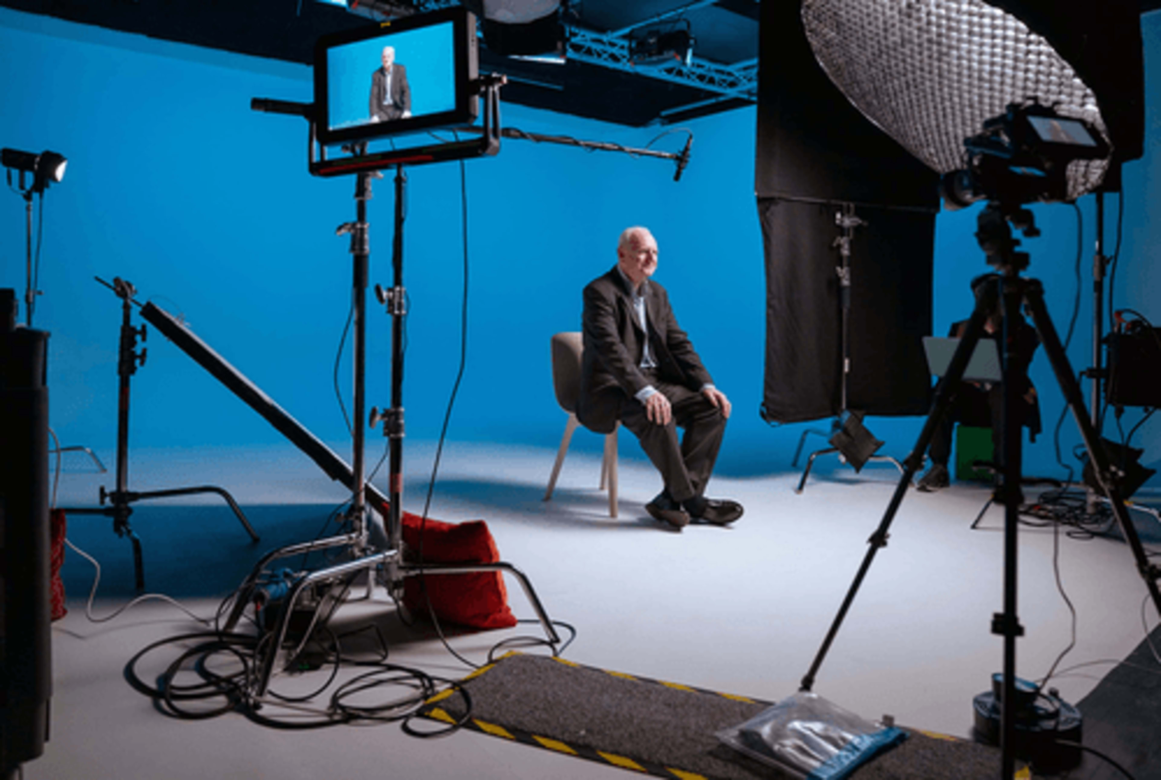 A man sits on a chair in a studio setup with blue backdrops, cameras, and lighting equipment surrounding him.