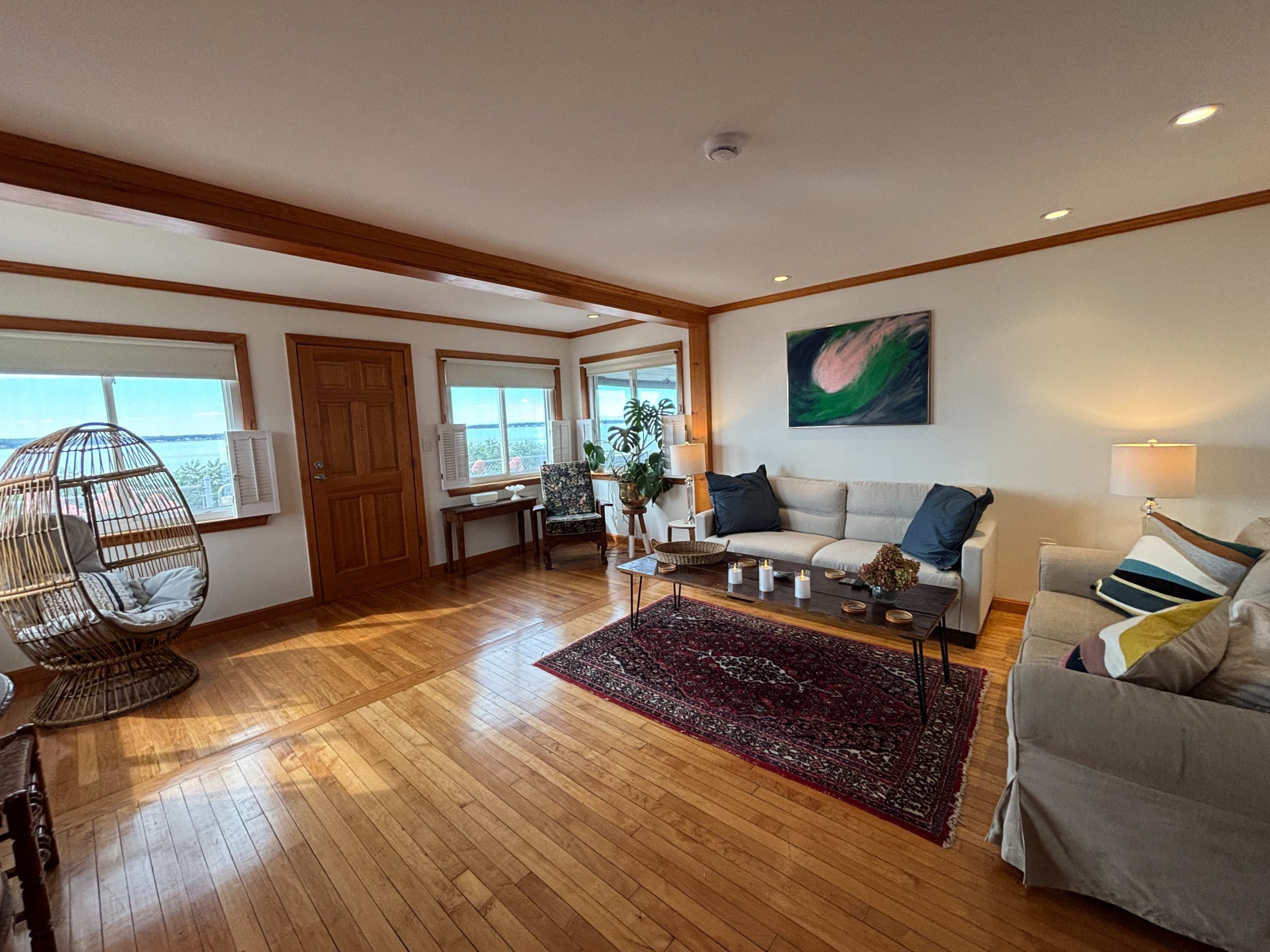 The image shows a bright living room with wooden floors, a cozy seating area featuring a couch and a chair, and a decorative rug in front of a coffee table.
