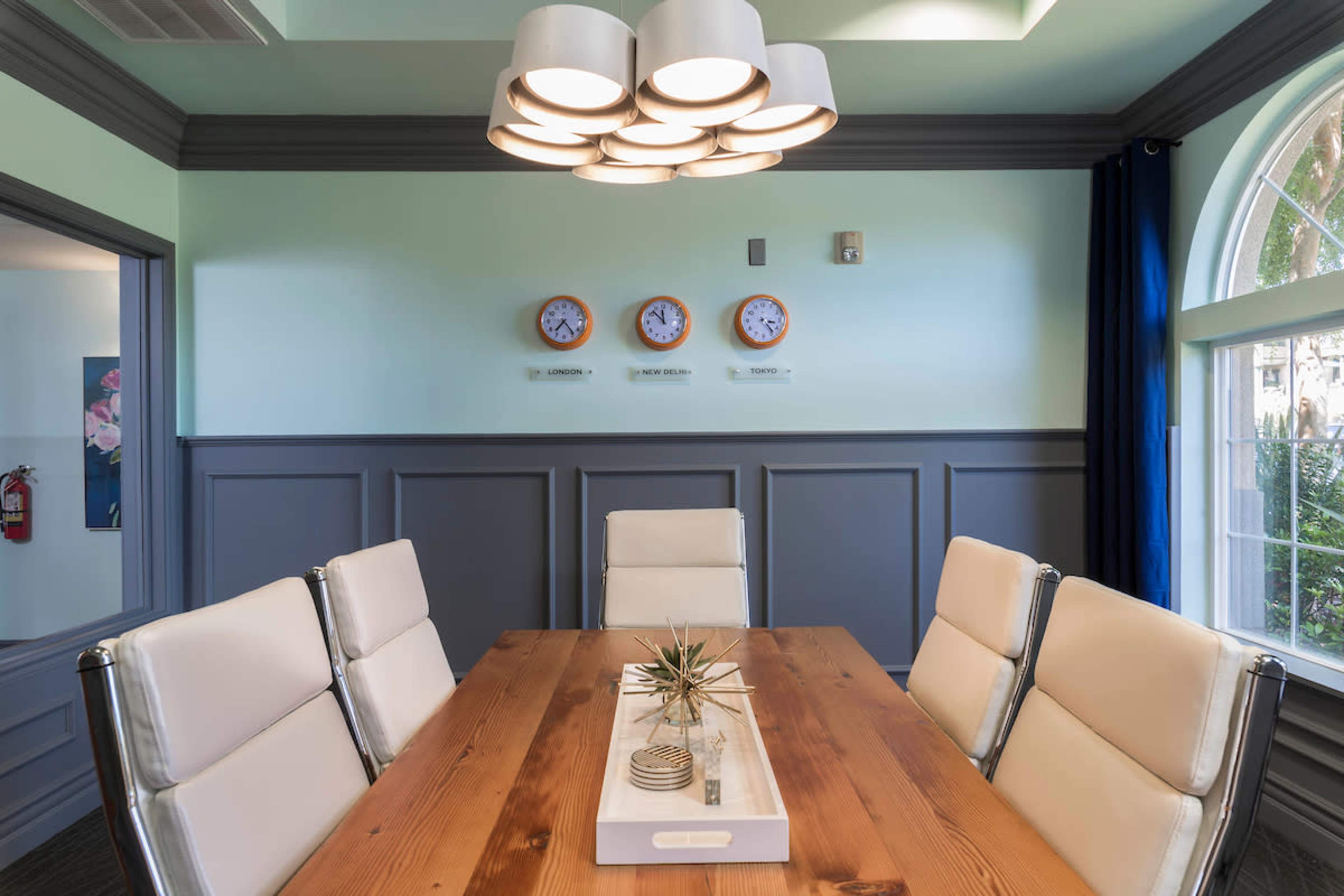 A modern conference room with a wooden table surrounded by white chairs, featuring decorative clocks on the wall and a large chandelier overhead.