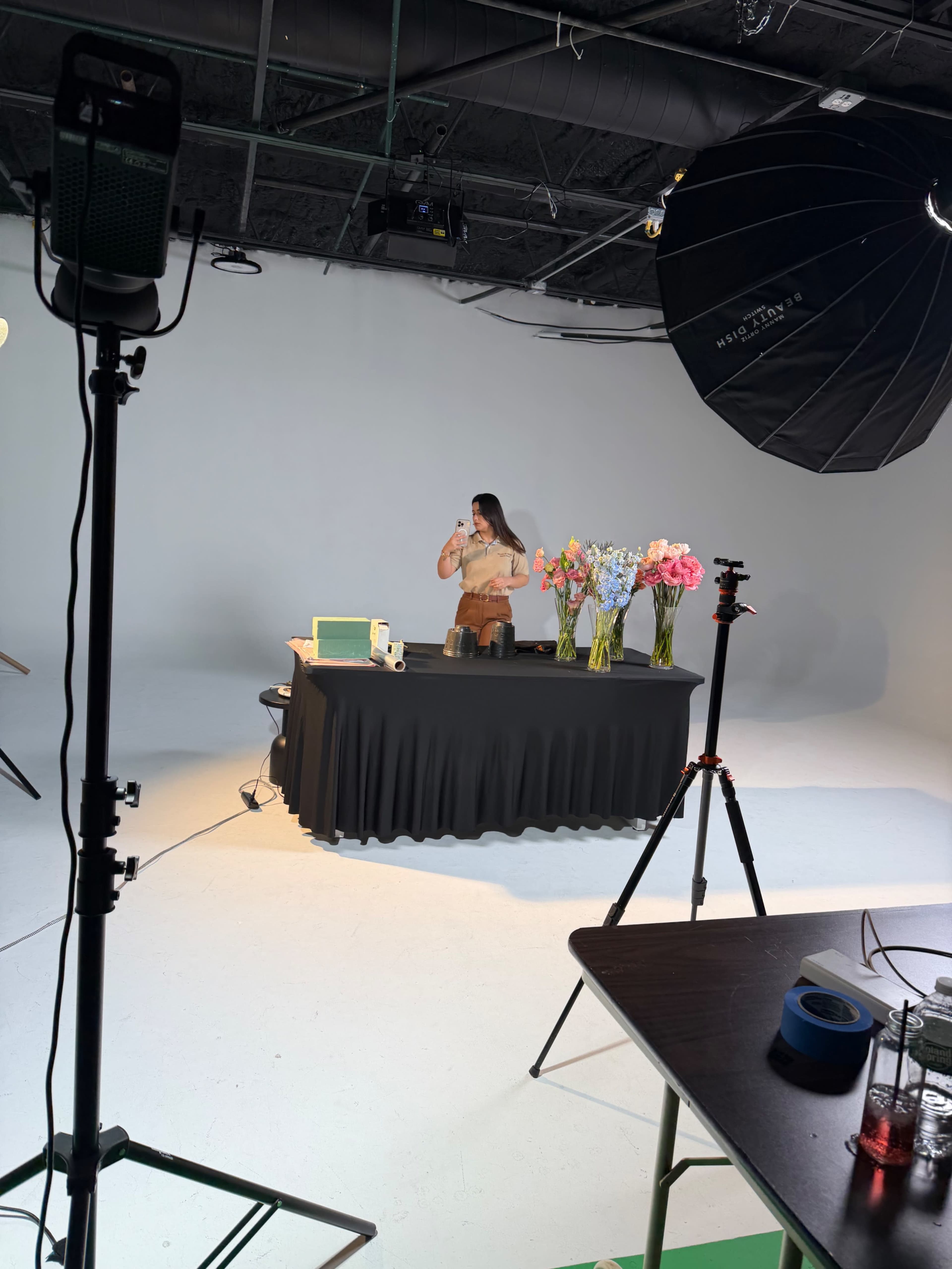 A woman is standing behind a table adorned with flowers and various items in a well-lit studio setting.