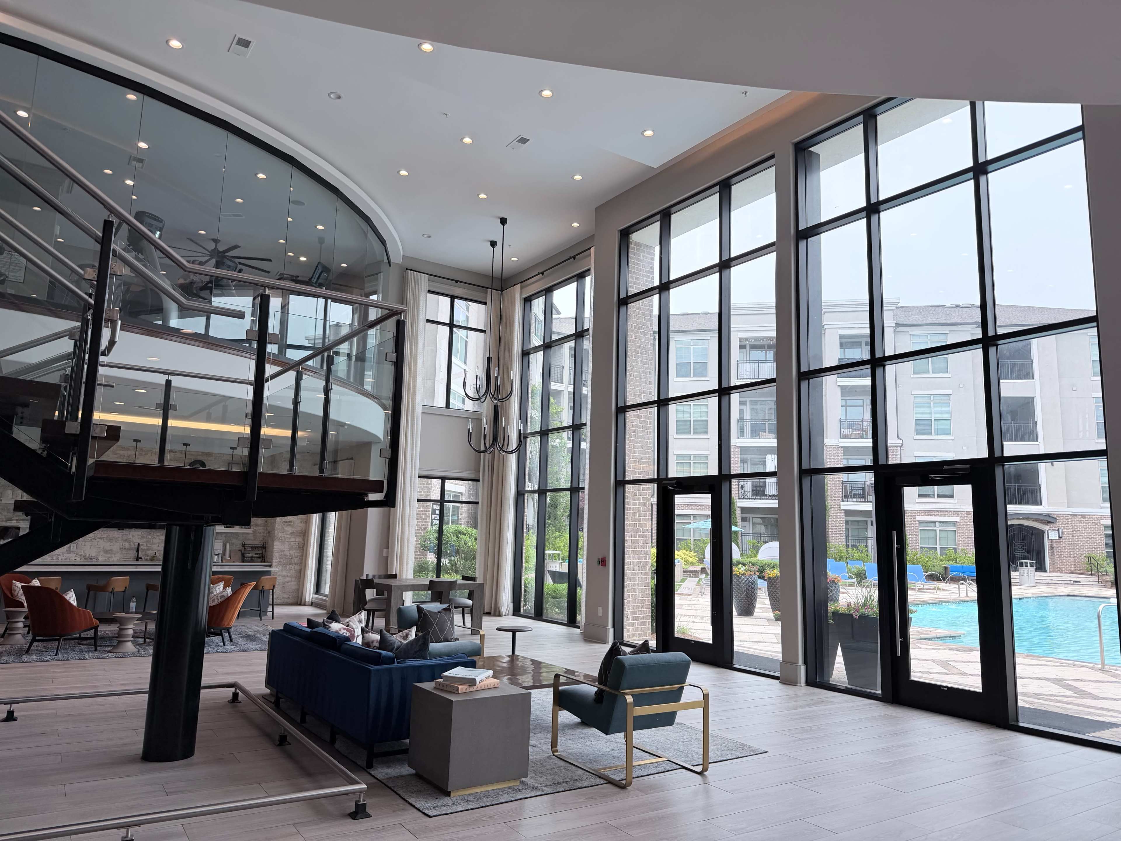 The image shows a modern lobby area with large windows overlooking a swimming pool, featuring stylish furniture and an open staircase.