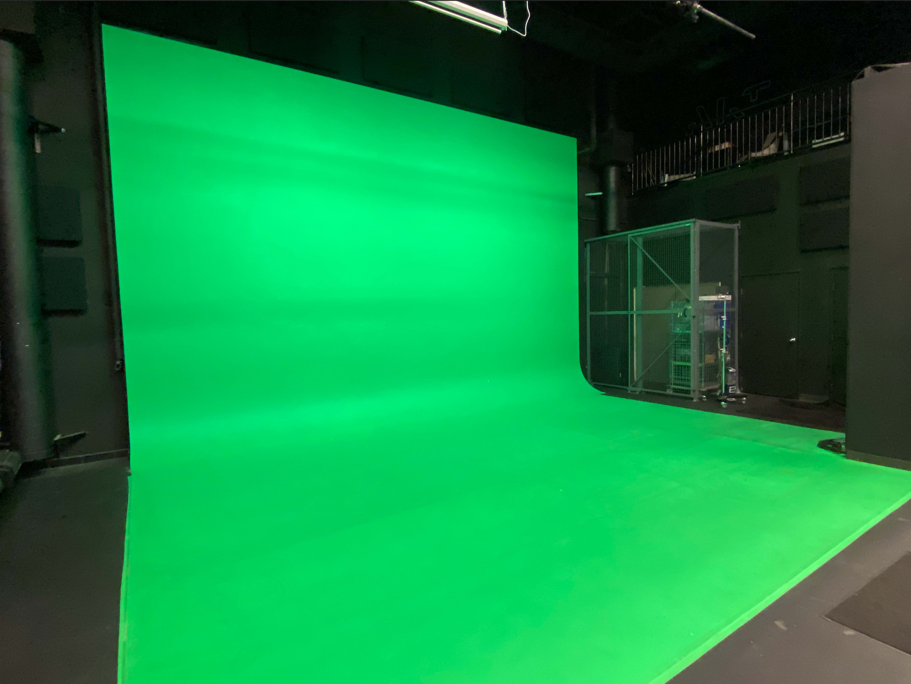 The image shows a green screen studio with a smooth green backdrop and a transparent enclosure in the corner.