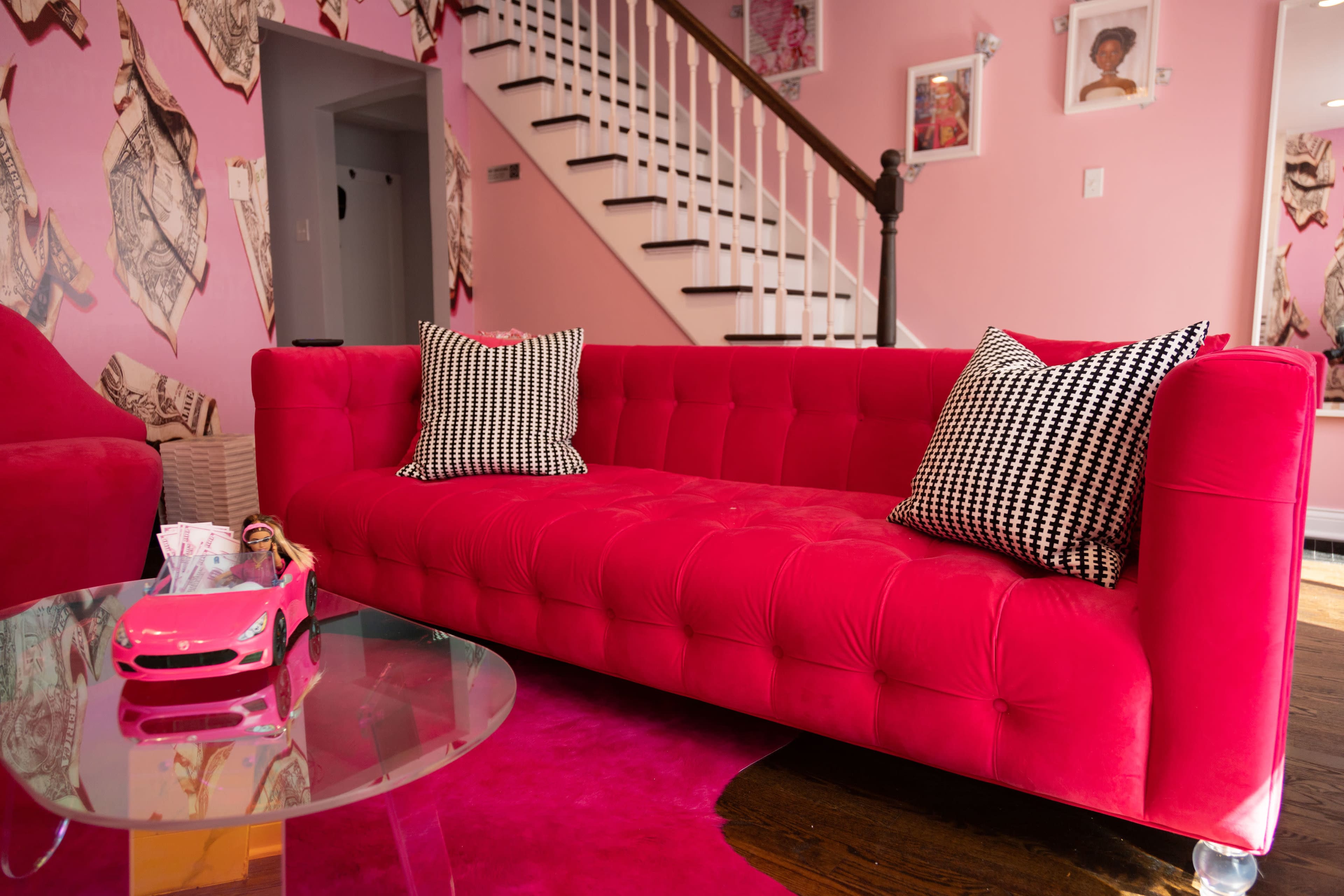 The scene features a bright pink velvet sofa with black and white checkered pillows, positioned next to a glass table and a toy car on a matching pink rug in a room decorated with pink walls and framed photos.