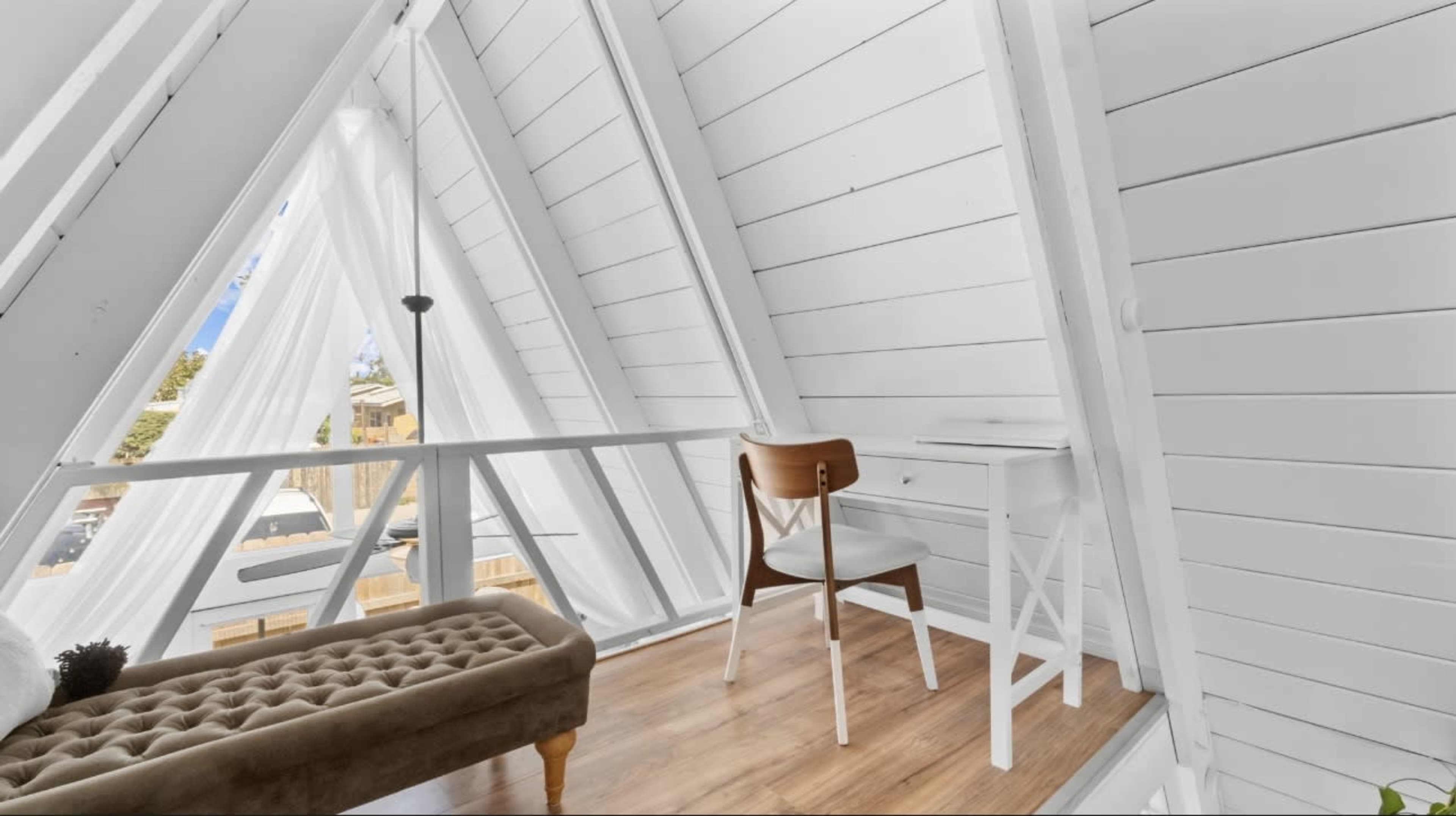 A bright, modern A-frame interior with white walls, a wooden floor, and a small desk and chair beside a window.
