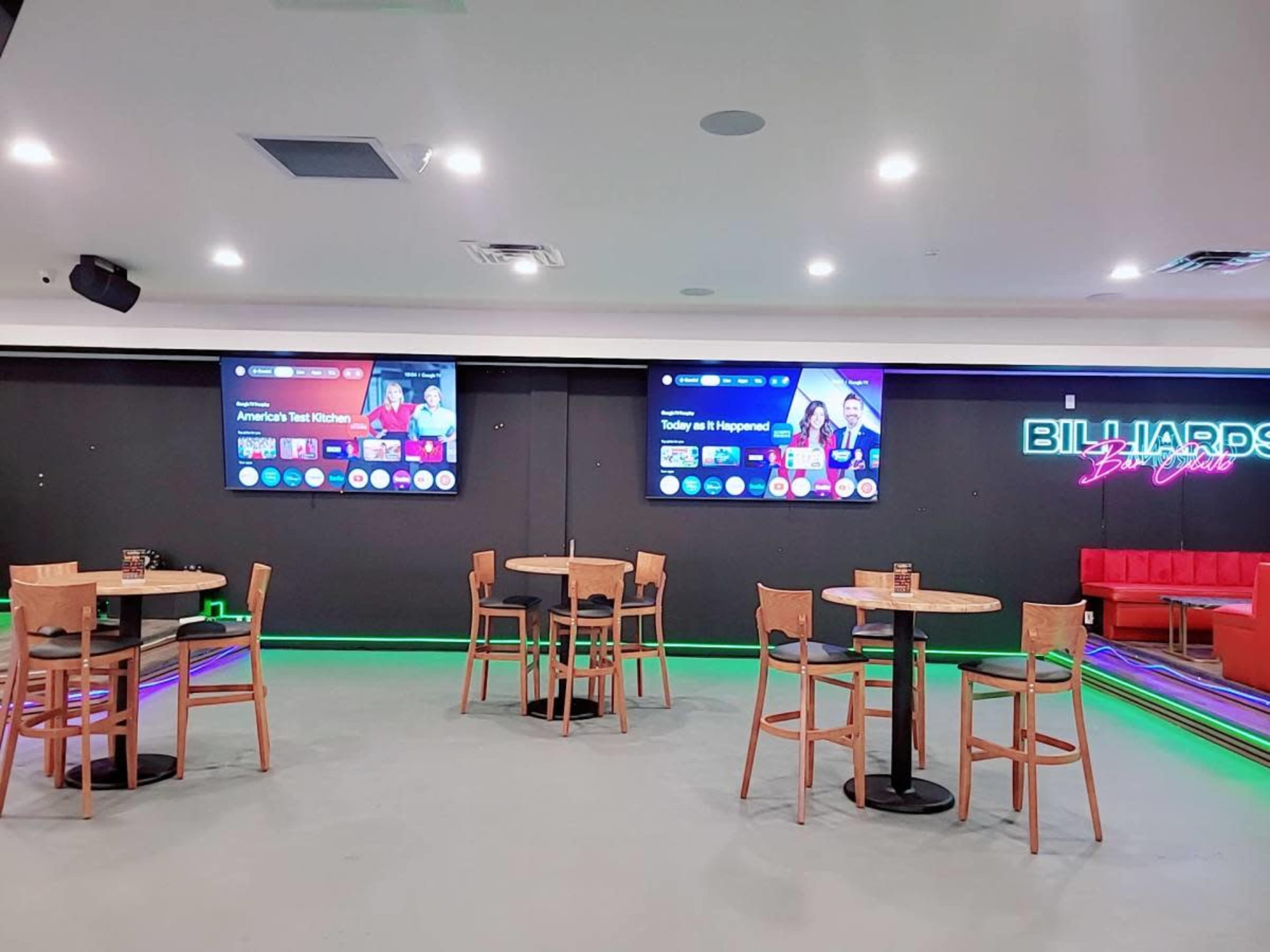 The image shows a modern recreational space with several wooden tables and chairs, large screens on the walls displaying sports content, and colorful LED lighting along the floor.