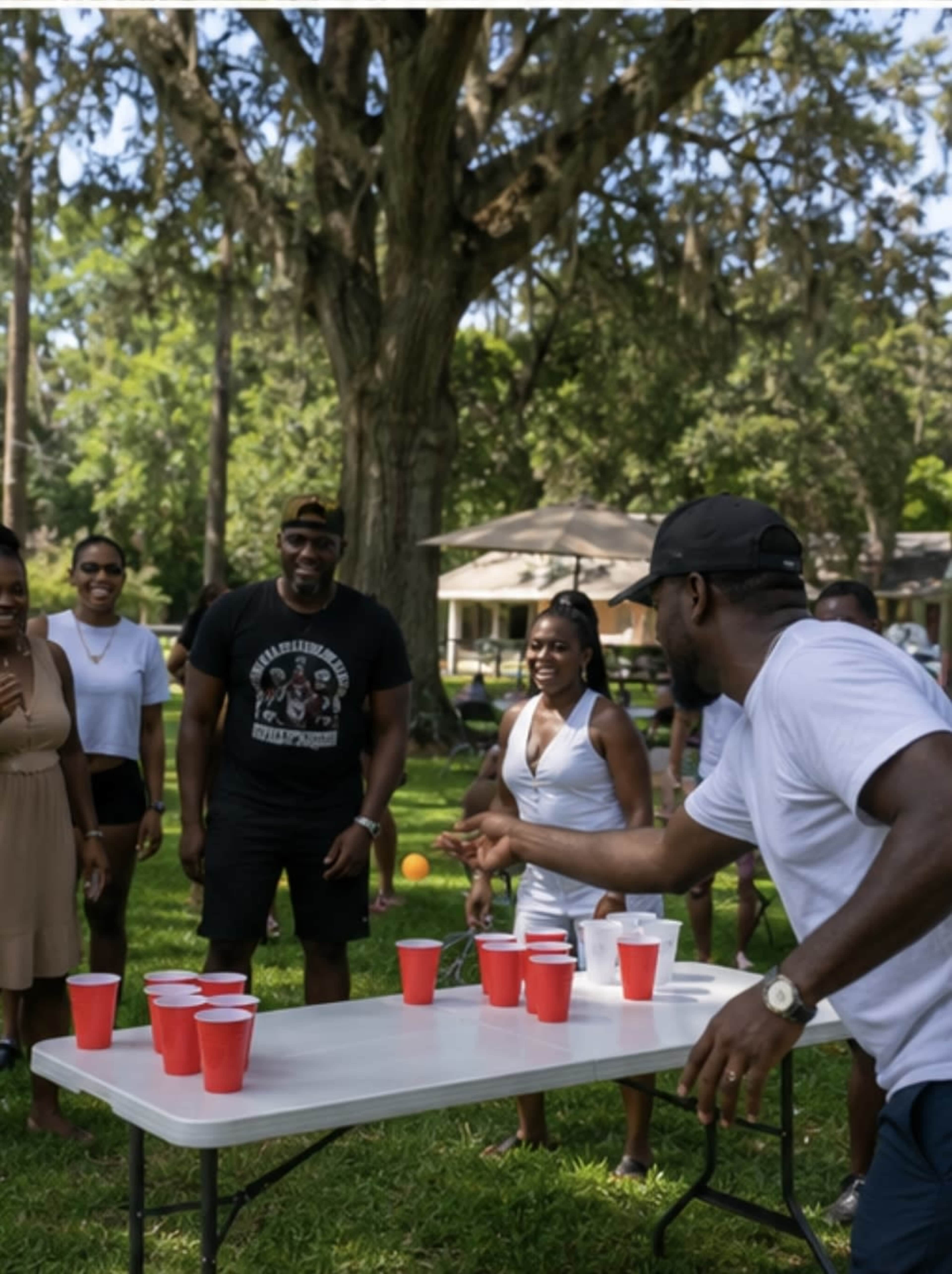 A group of people gathers around a table in a park, watching as a man throws a ball towards a set of red cups.