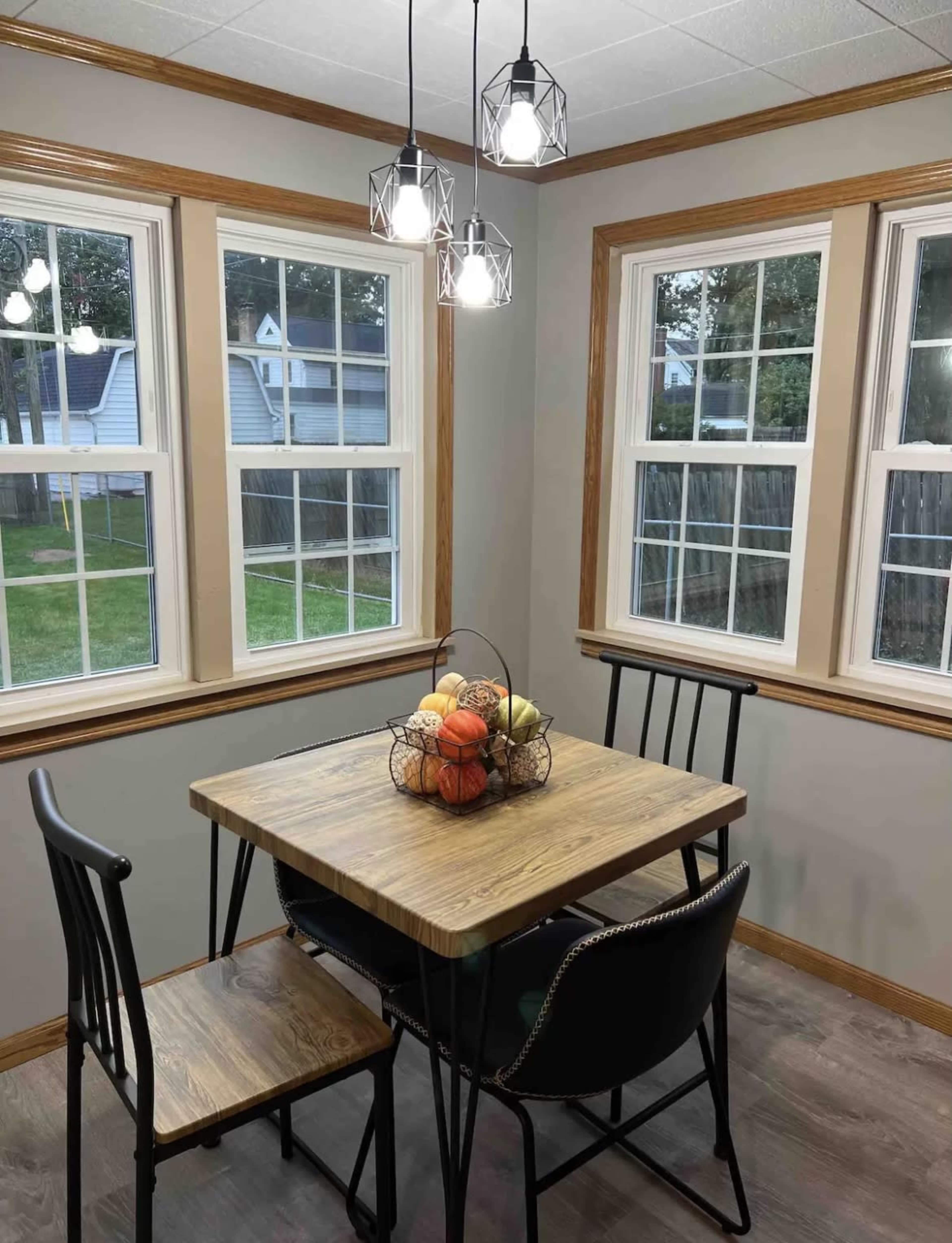 A small dining area features a wooden table with a fruit centerpiece and black chairs near windows.