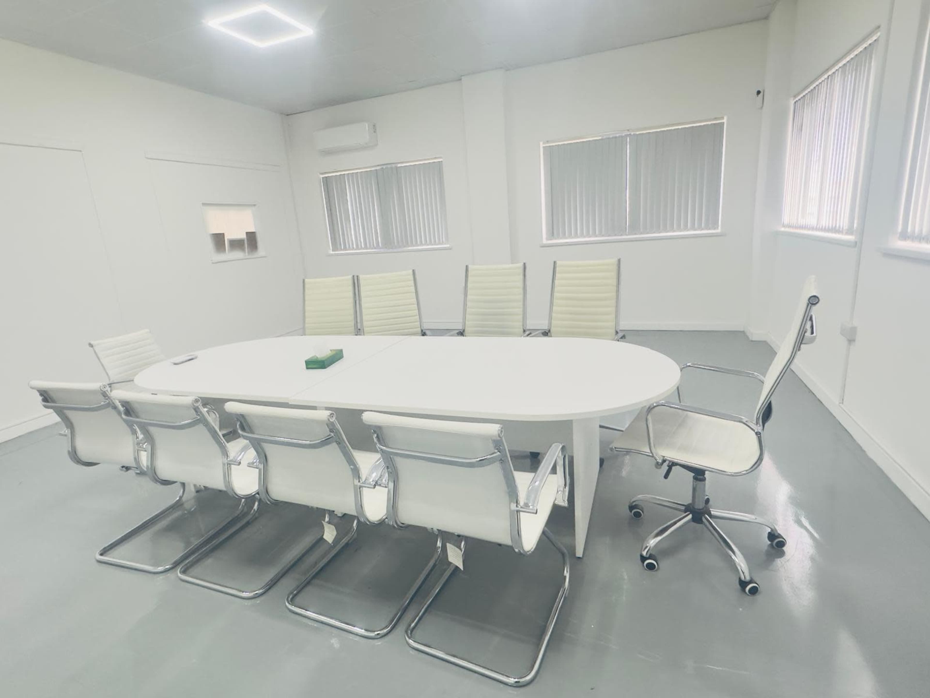 A white conference room features an oval table surrounded by six white chairs and a rolling chair, with large windows allowing natural light.